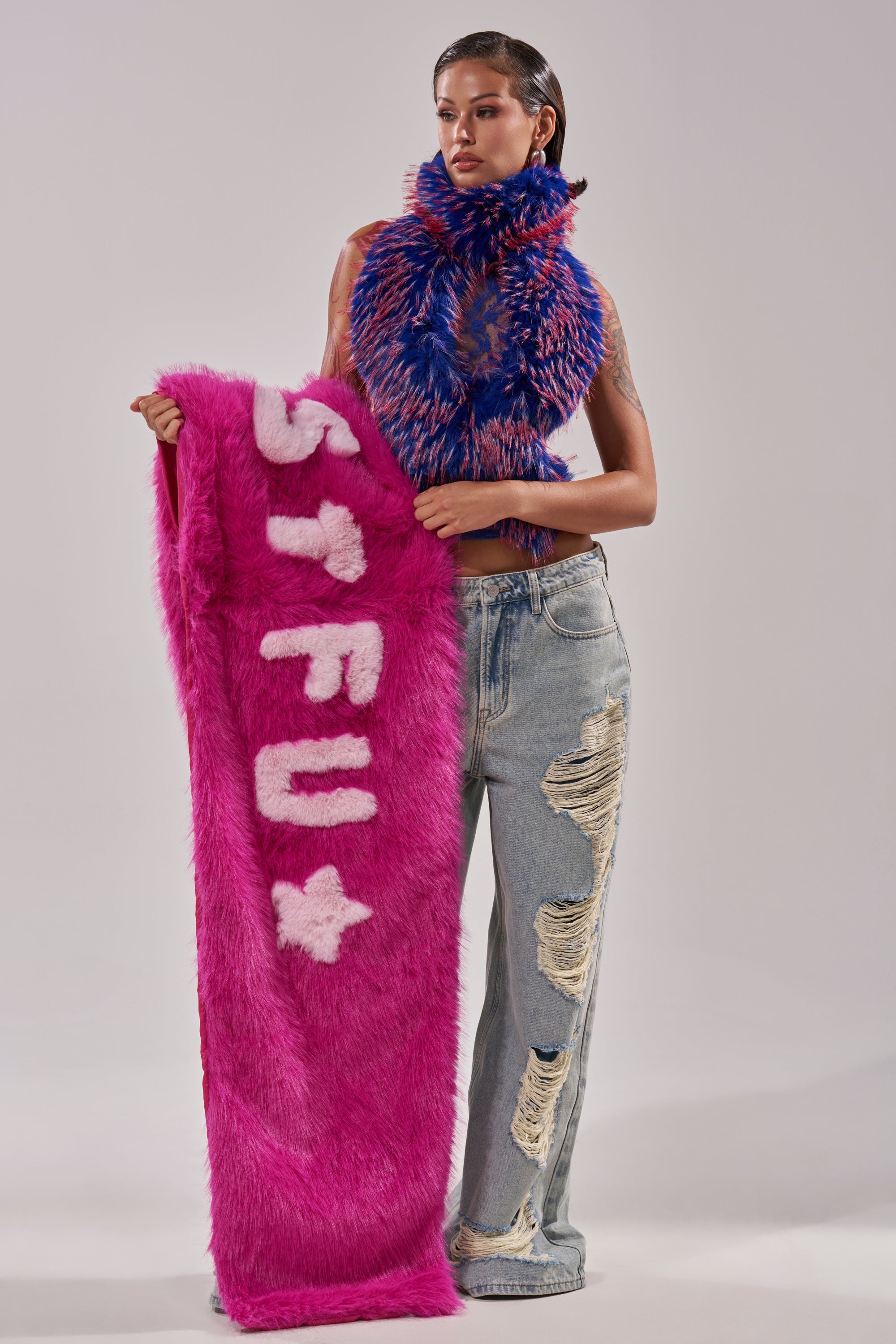 Wearing distressed jeans, a person holds a large pink furry "STFU" sign with a heart, styled with the colorful and oversized STFU FAUX FUR SCARF, against a plain background. 1