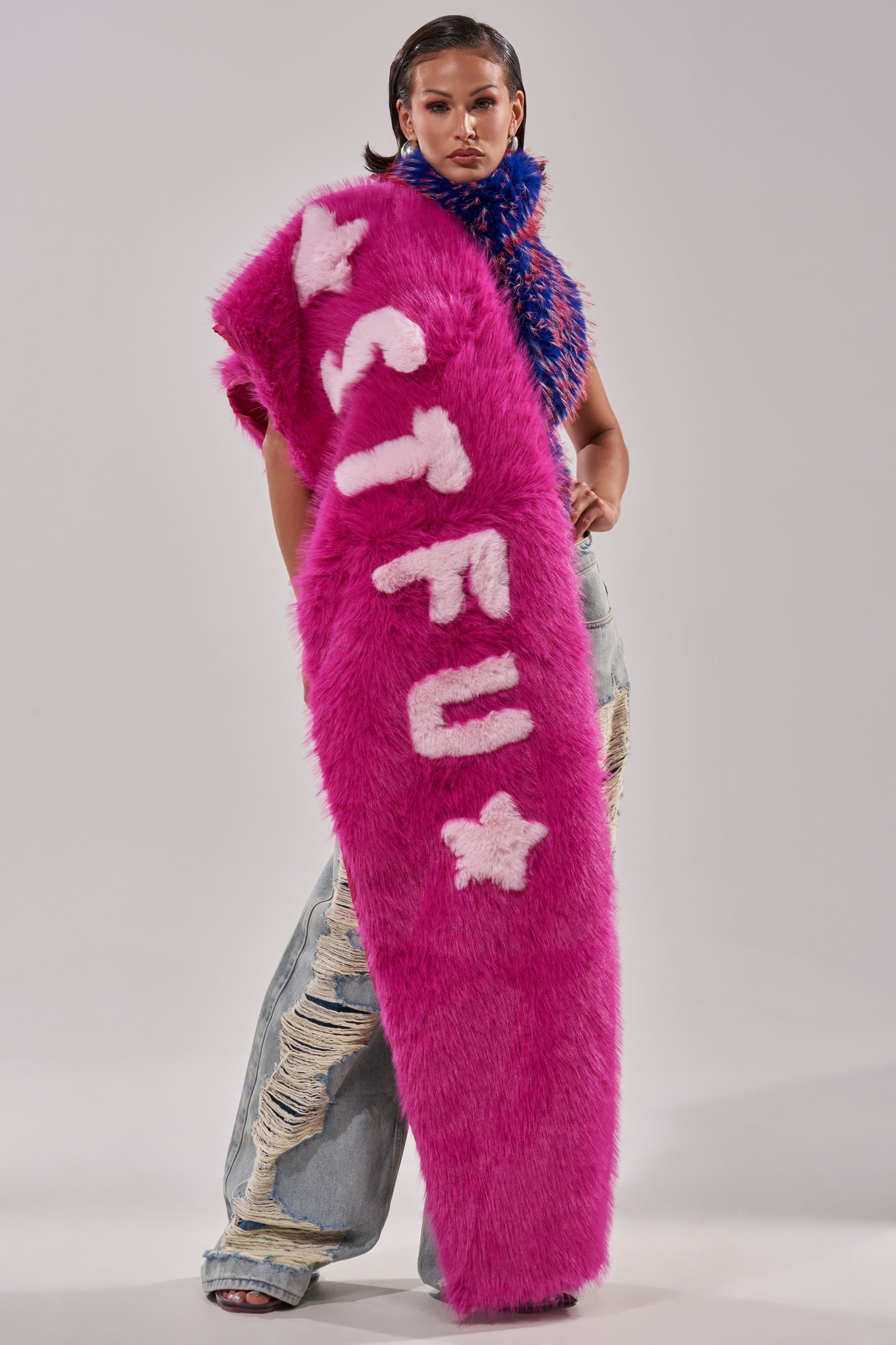 A model wears distressed jeans and the oversized STFU FAUX FUR SCARF with “STFU” and white stars, paired with a blue and pink textured top, against a plain light background. 2