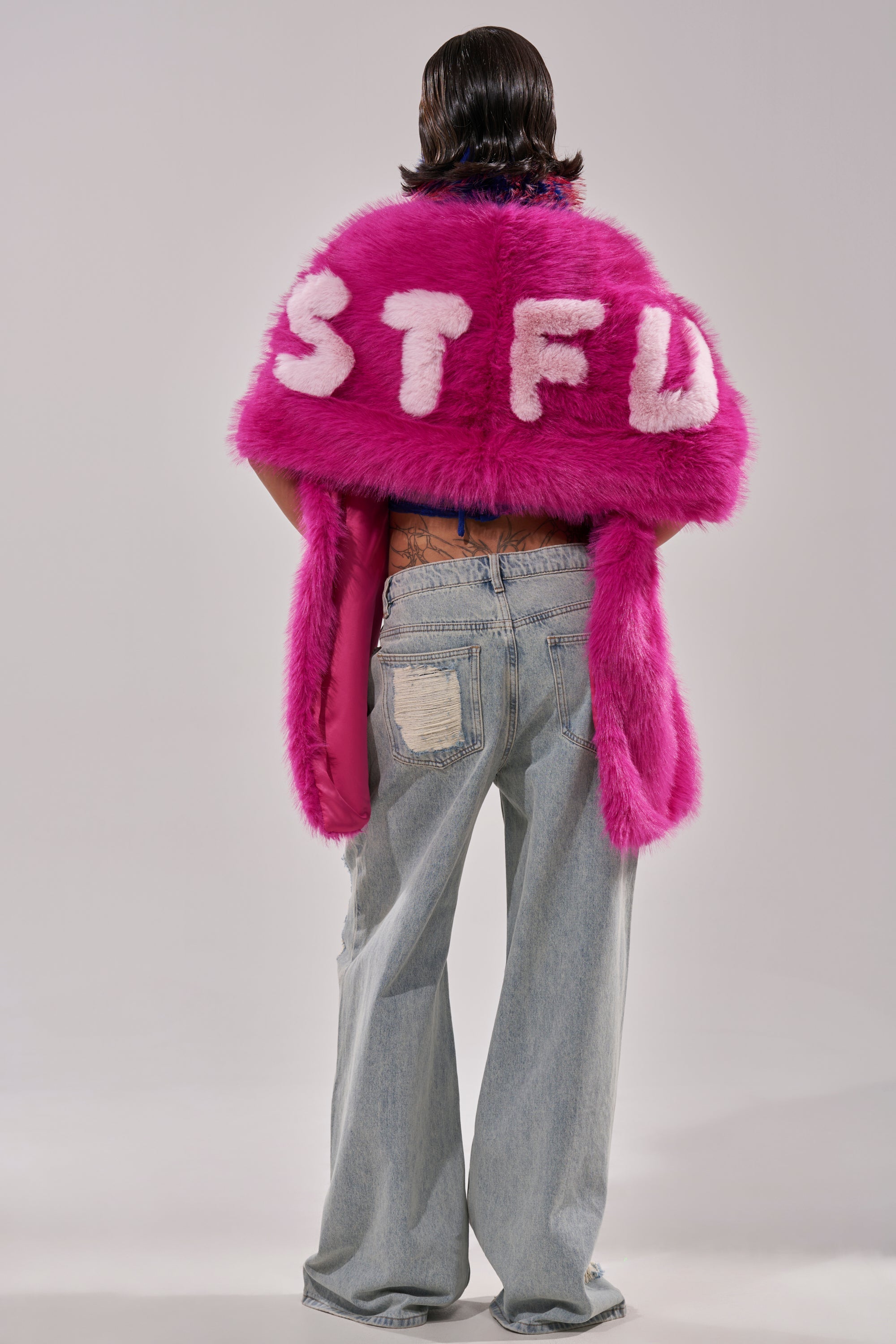 A person with short dark hair models the STFU FAUX FUR SCARF, a bright pink accessory featuring bold white "STFU" lettering on the back. They pair it with light blue baggy jeans and stand against a plain, light background. 4