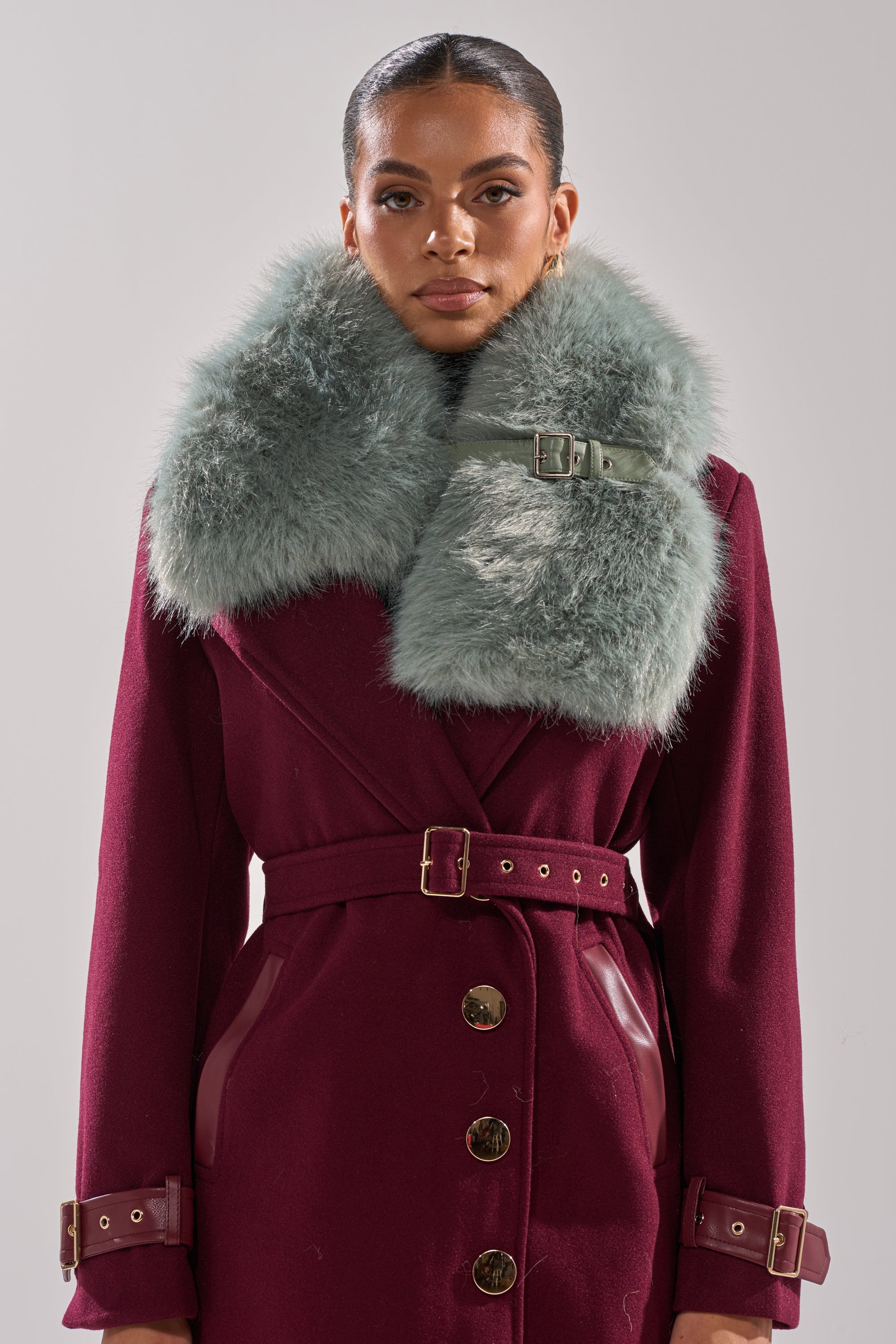 A woman stands facing forward in the HAVE IT ALL FAUX FUR SHAWL: a deep burgundy coat with gold buttons, wide belt, and a large pale green faux fur shawl collar secured by a buckle, set against a plain light gray background. 0