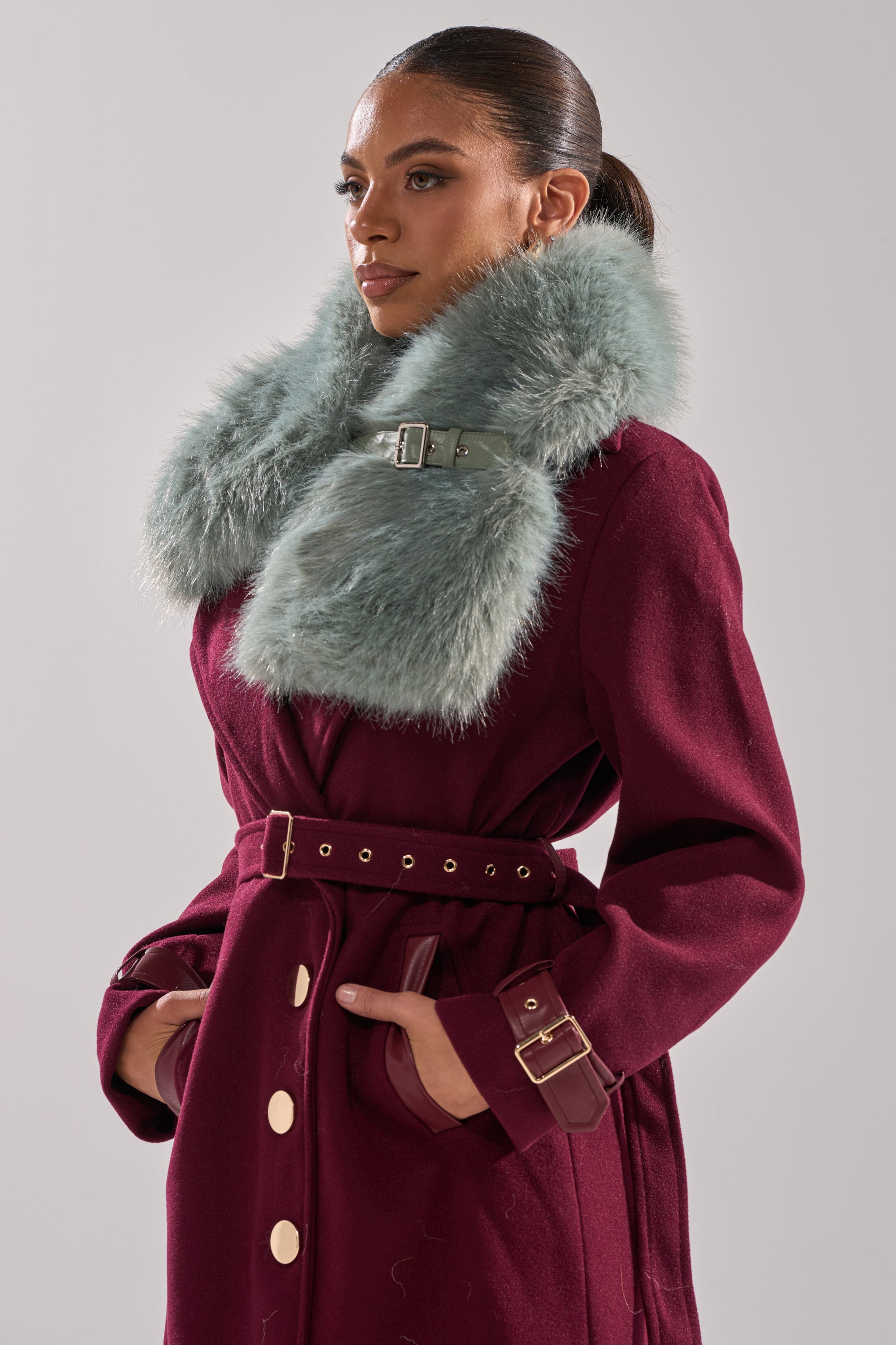 A woman wears the HAVE IT ALL FAUX FUR SHAWL—an eye-catching, large, fluffy light blue accessory with a buckle—over a burgundy coat with gold buttons and a belt. Her hands are in her pockets and her hair is in a sleek ponytail. 1