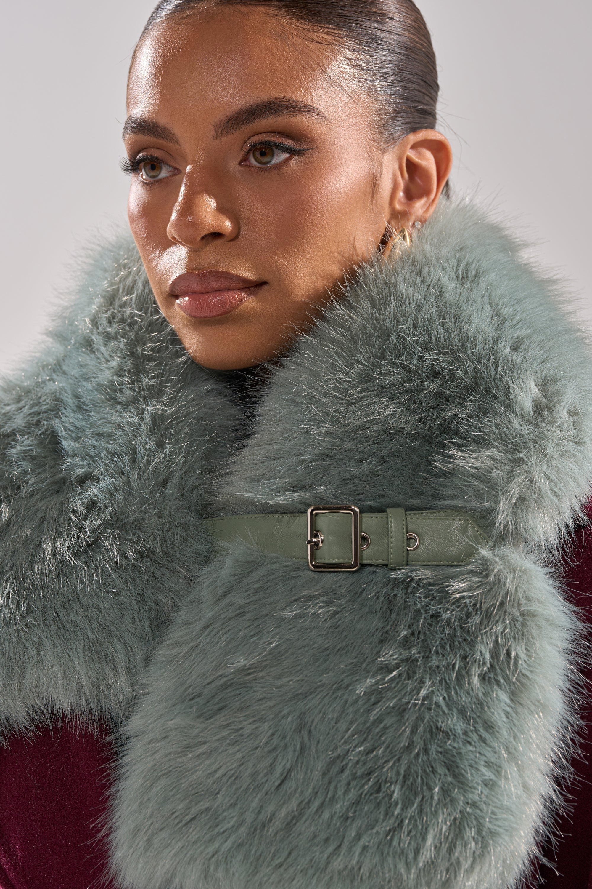 A woman with slicked-back hair wears the HAVE IT ALL FAUX FUR SHAWL, a light green piece with a leather buckle over a burgundy top, gazing softly to the side—an elegant accessory for eveningwear. 2
