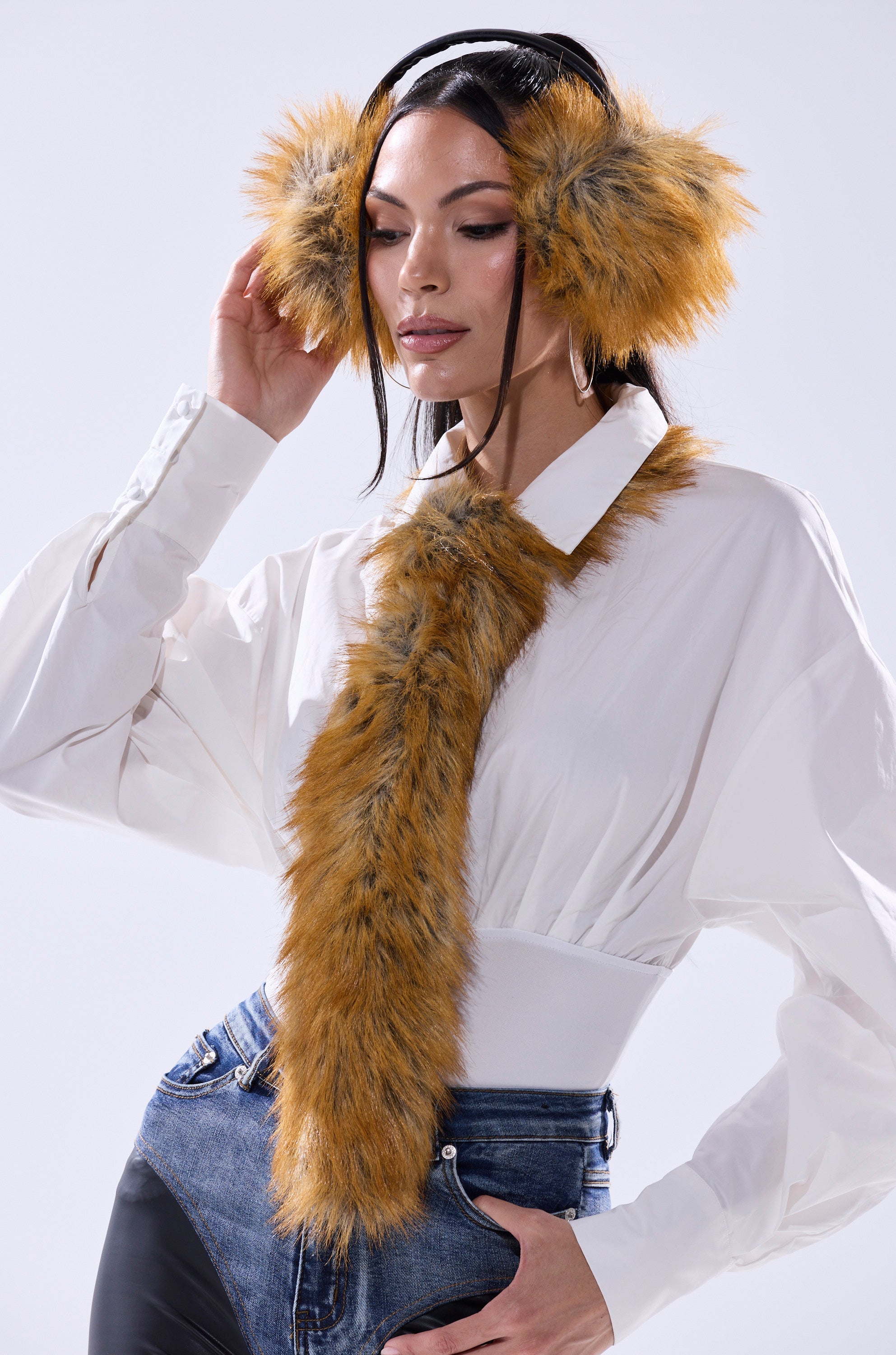 A woman wears the REALLY LIKE THAT FAUX FUR TIE in plush orange-brown as earmuffs and a matching scarf over a white puff-sleeve blouse and high-waisted blue jeans, posing with one hand touching an earmuff and looking slightly down. 0