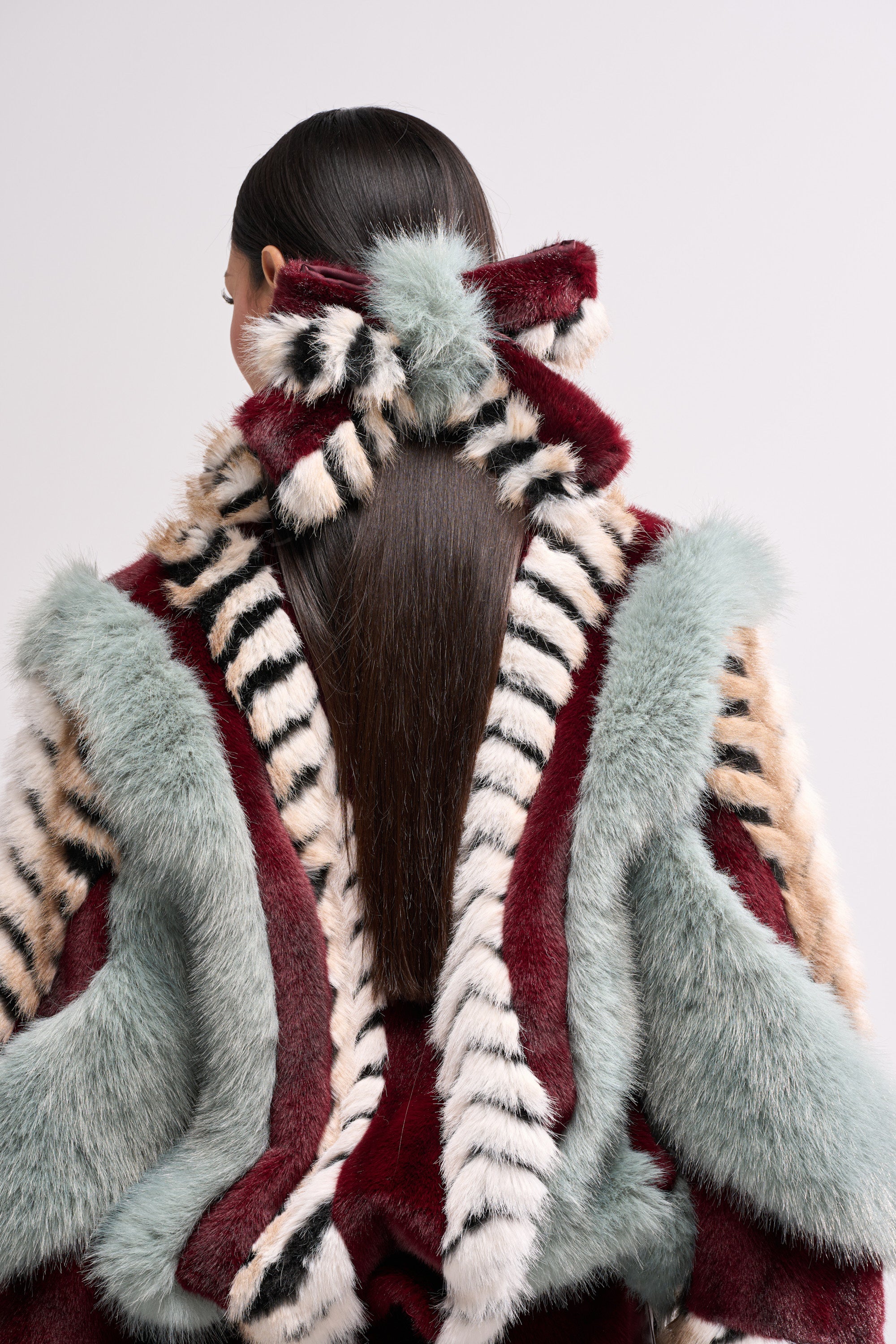 A person with long, straight brown hair wears a bow-shaped HAVE IT ALL FAUX FUR HAIR CLIP and a striped fur coat in burgundy, pale blue, cream, and black, standing with their back to the camera.