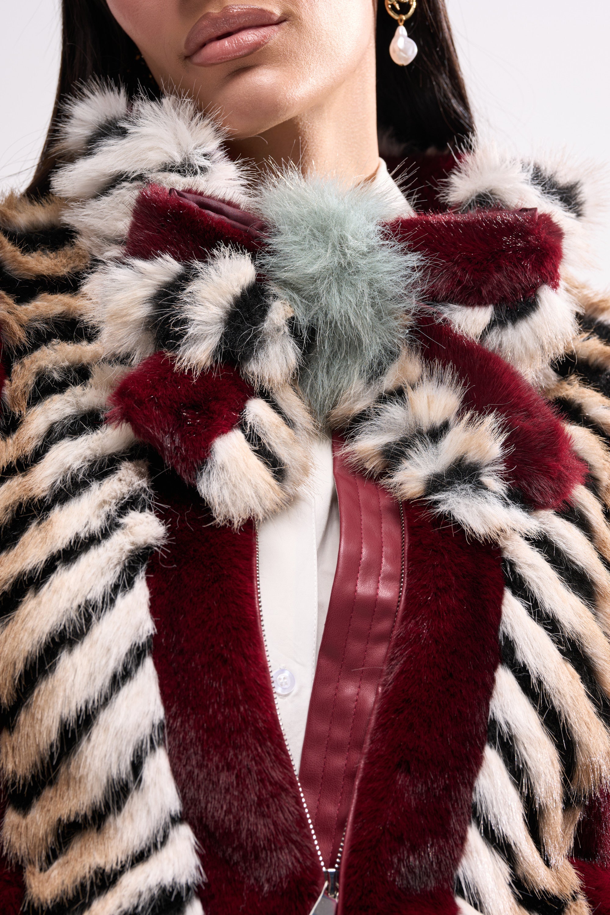 A woman shows her lower face and upper body in a luxurious maroon, black, and white striped faux fur coat with a large bow collar, blue pompom, gold pearl earring, and the HAVE IT ALL FAUX FUR HAIR CLIP in her hair. 1