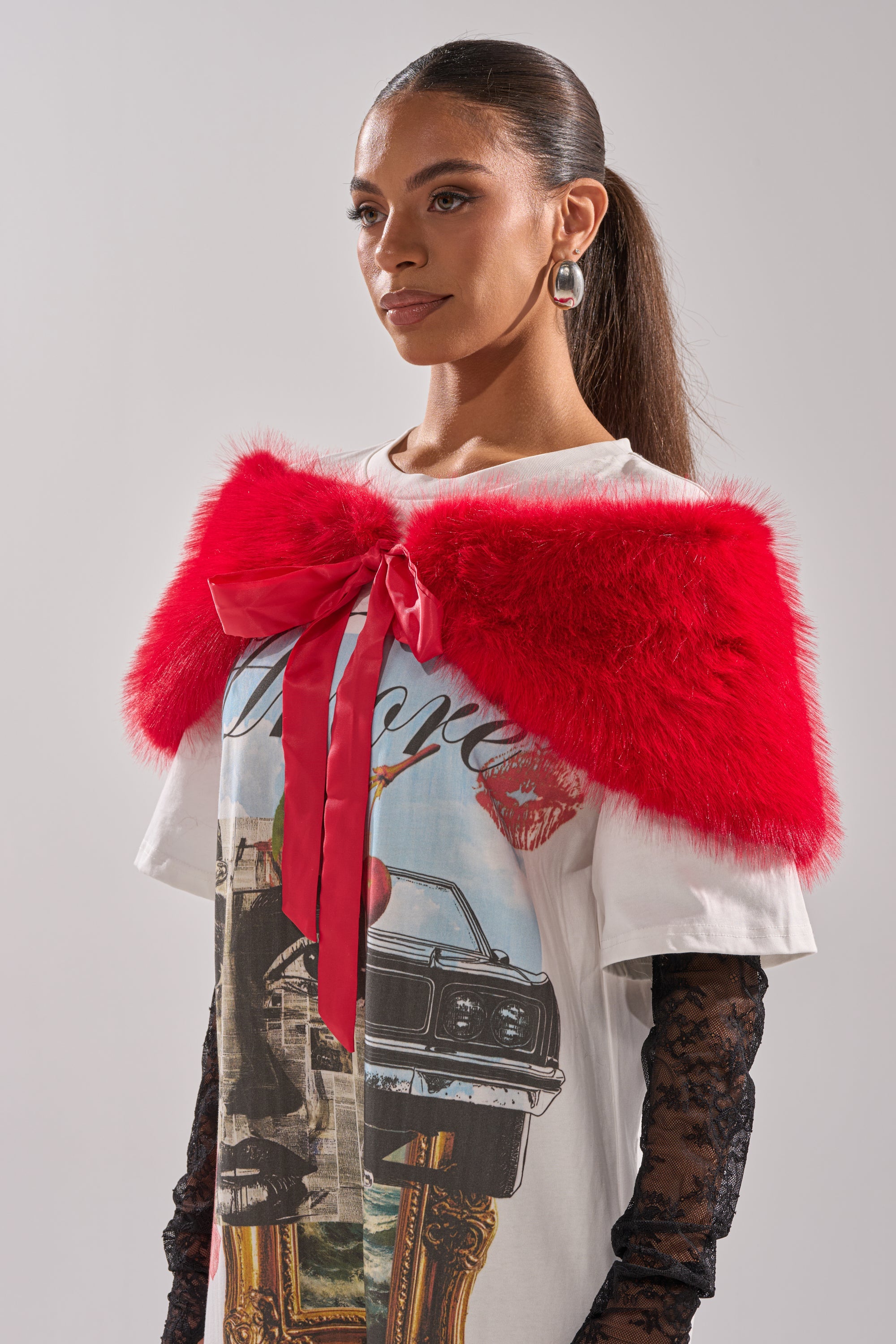 A woman with slicked-back hair models the WITH A BOW FAUX FUR SHAWL in bright red, featuring a ribbon detail, styled over a graphic T-shirt and lace sleeves against a plain light background. 2