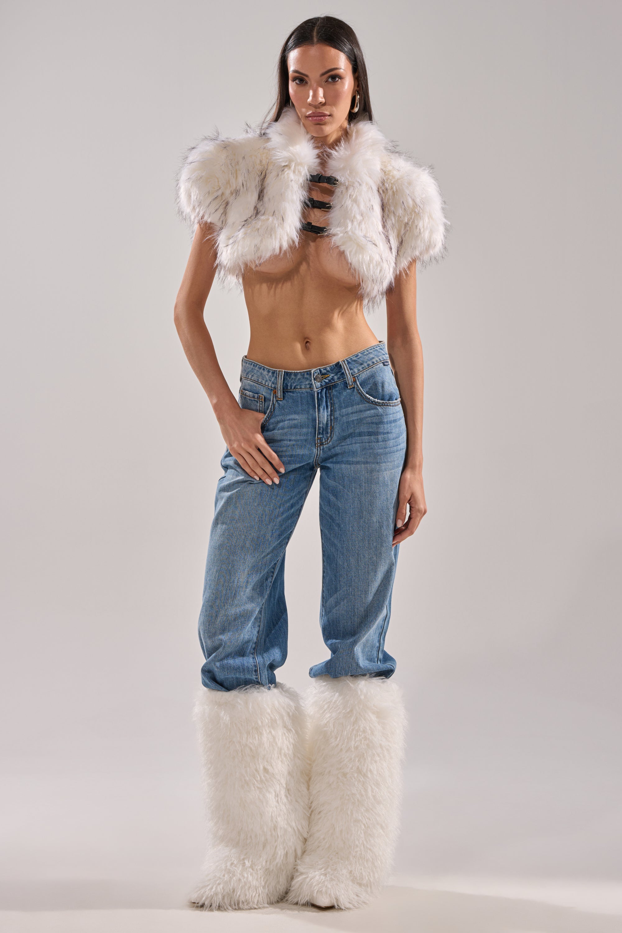 A woman poses confidently against a light background in blue jeans and the LUXE LIFE FAUX FUR SHAWL IN WHITE, styled cropped to reveal her midriff. She completes the look with matching white furry boots. 4