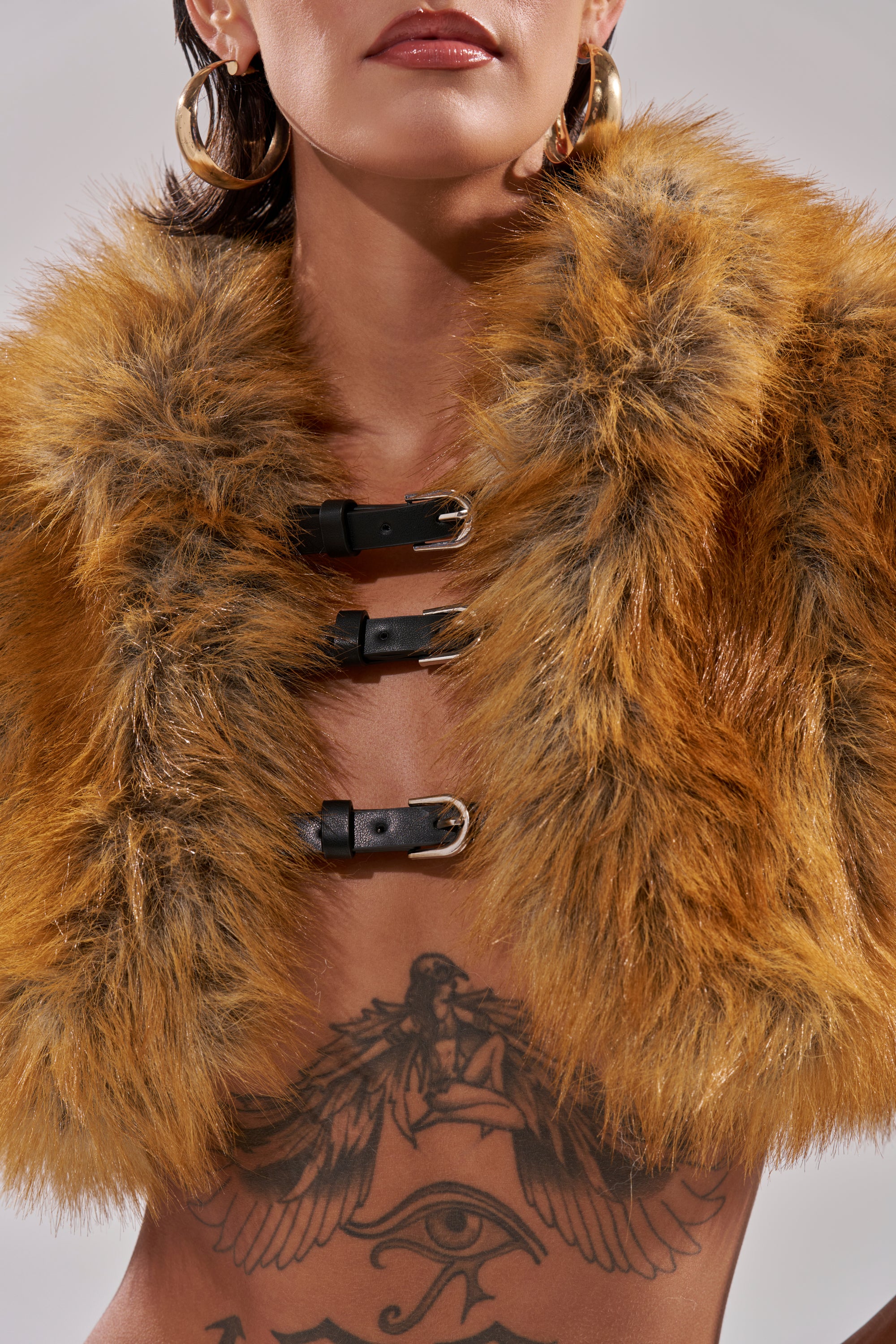 A person wears the LUXE LIFE FAUX FUR SHAWL IN BROWN, accented with large hoop earrings and showcasing a striking chest tattoo of a winged figure and an Eye of Horus for an intriguing, bold look.