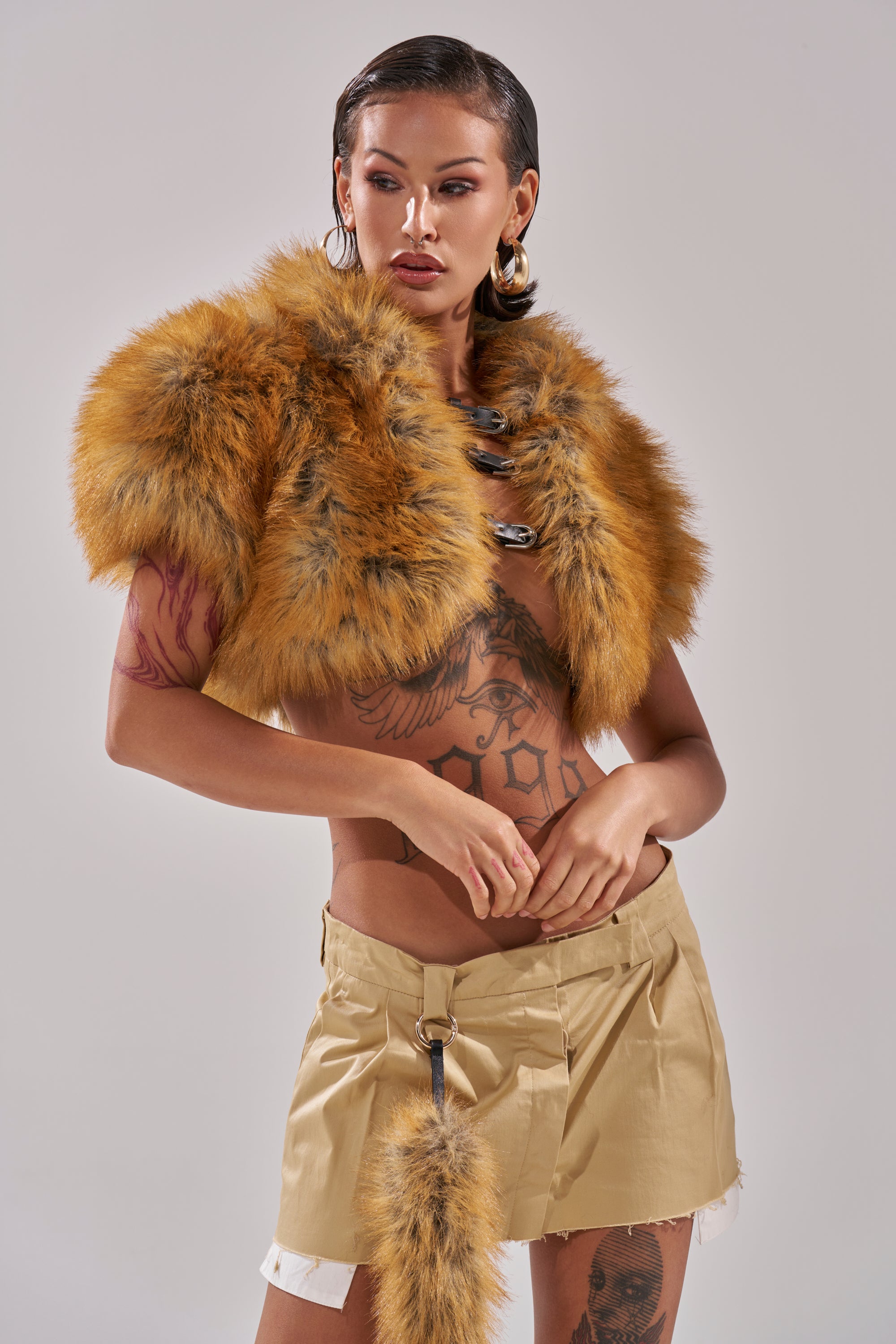 A woman with slicked-back hair wears the LUXE LIFE FAUX FUR SHAWL IN BROWN and a beige mini skirt with a fur tail accessory, showcasing tattoos on her torso and arms against a light background.