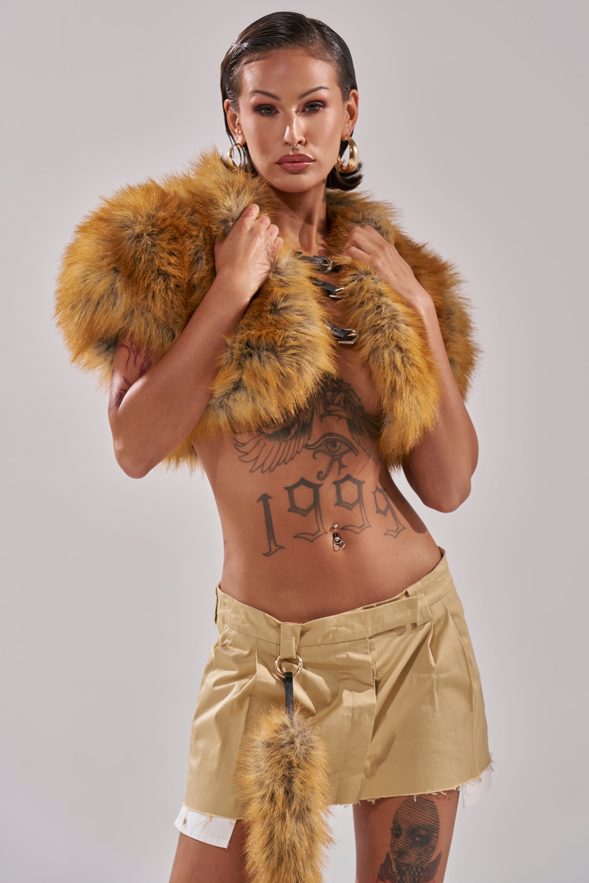 A person with slicked-back hair wears gold hoop earrings, beige shorts, a fur tail accessory, and the LUXE LIFE FAUX FUR SHAWL IN BROWN, showing tattoos on their chest and legs against a light gray background.