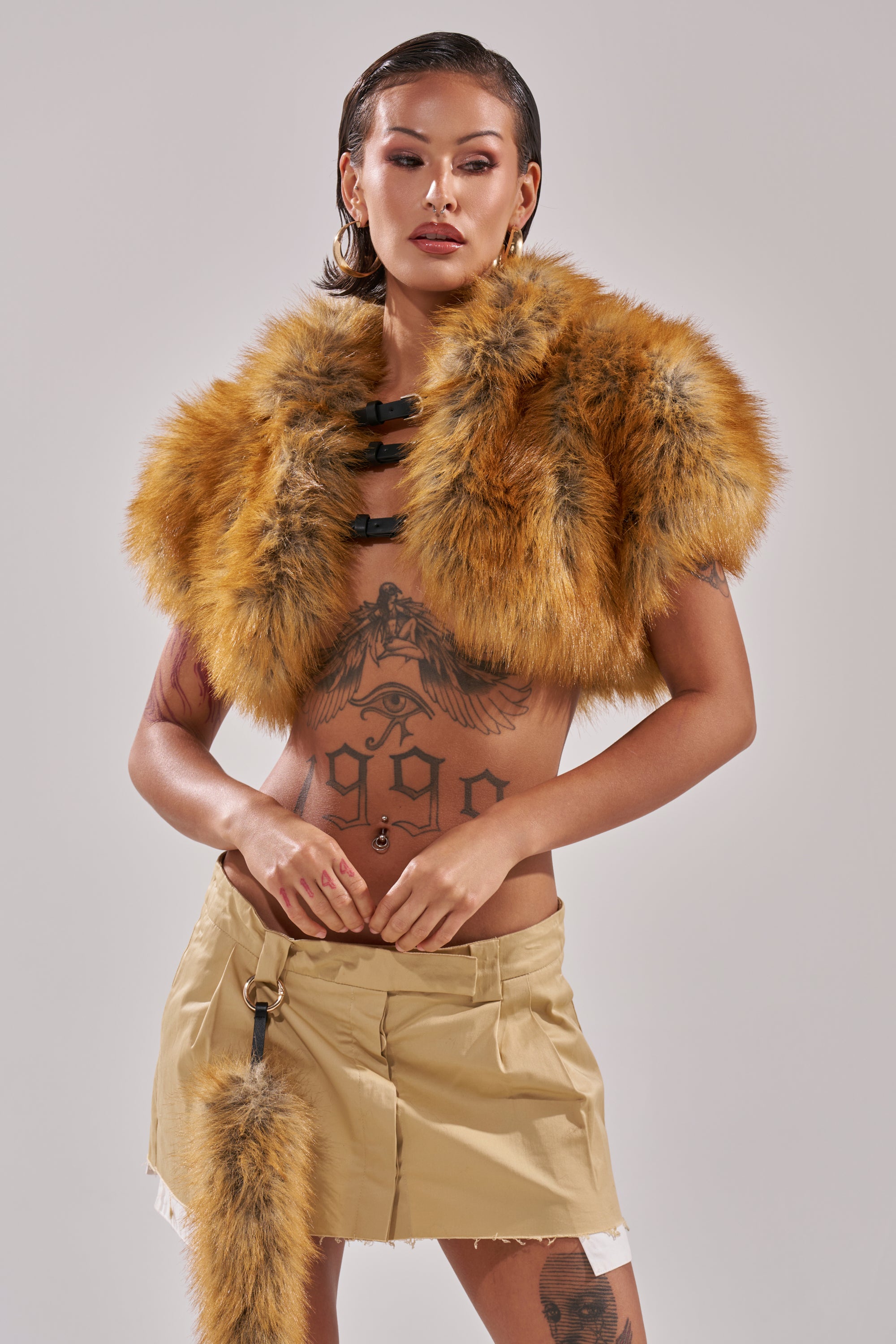 A tattooed person with slicked-back hair poses against a plain background, wearing the LUXE LIFE FAUX FUR SHAWL IN BROWN styled with a beige mini skirt and black fasteners.