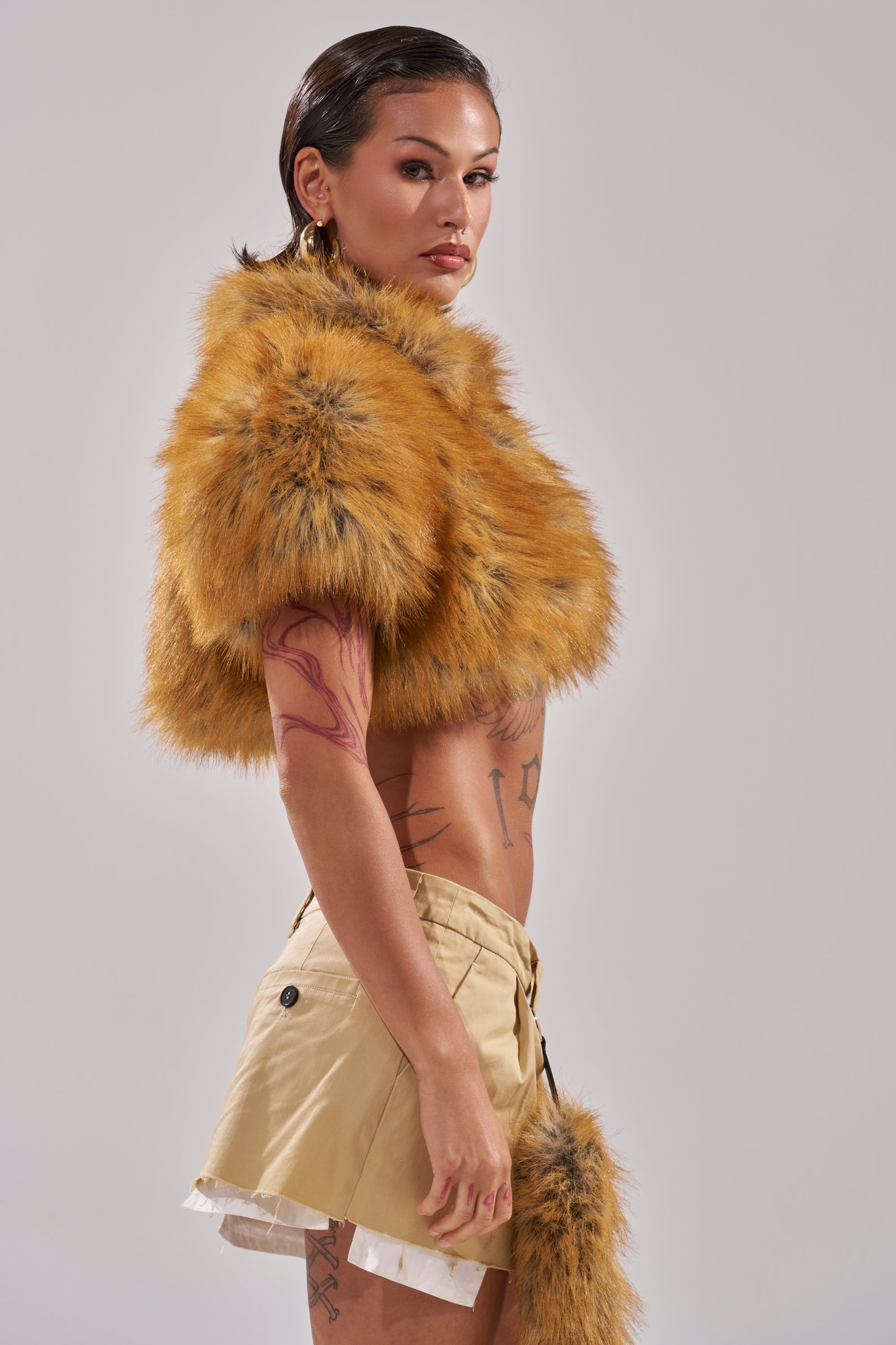 A person with slicked-back dark hair and visible tattoos stands sideways against a plain backdrop, wearing the LUXE LIFE FAUX FUR SHAWL IN BROWN, a tan mini skirt, and gold hoop earrings.