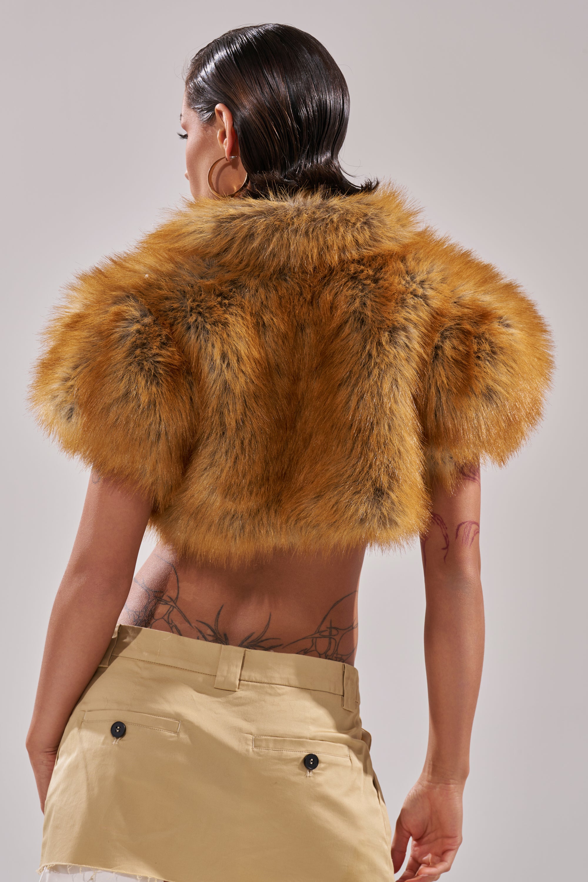 A person with wet, slicked-back hair is shown from the back, displaying side and lower-back tattoos while wearing the LUXE LIFE FAUX FUR SHAWL IN BROWN and beige pants.