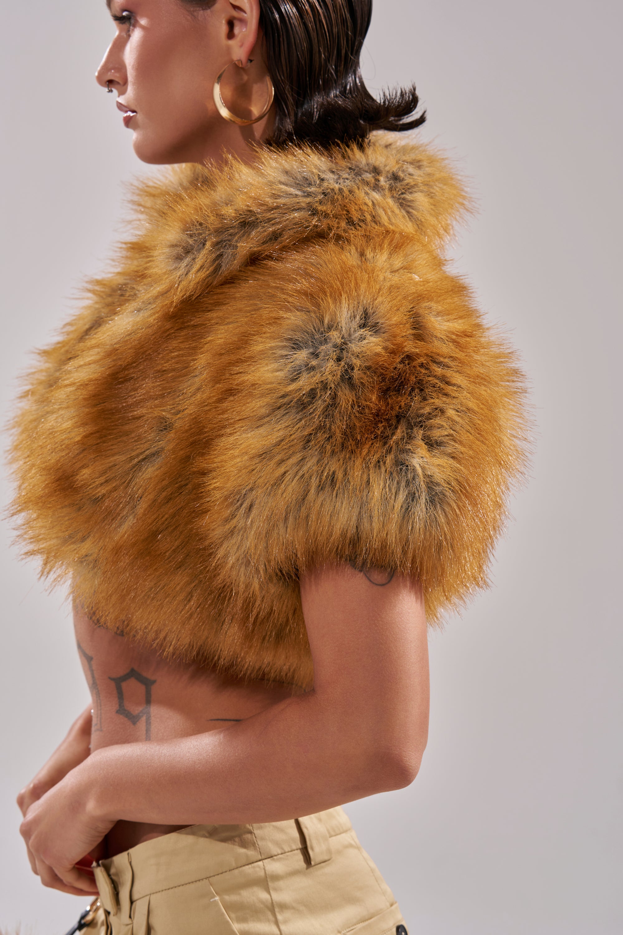 A person with slicked-back hair wears large hoop earrings, beige pants, and the LUXE LIFE FAUX FUR SHAWL IN BROWN, revealing a “19” tattoo on their torso against a plain, light-colored background.