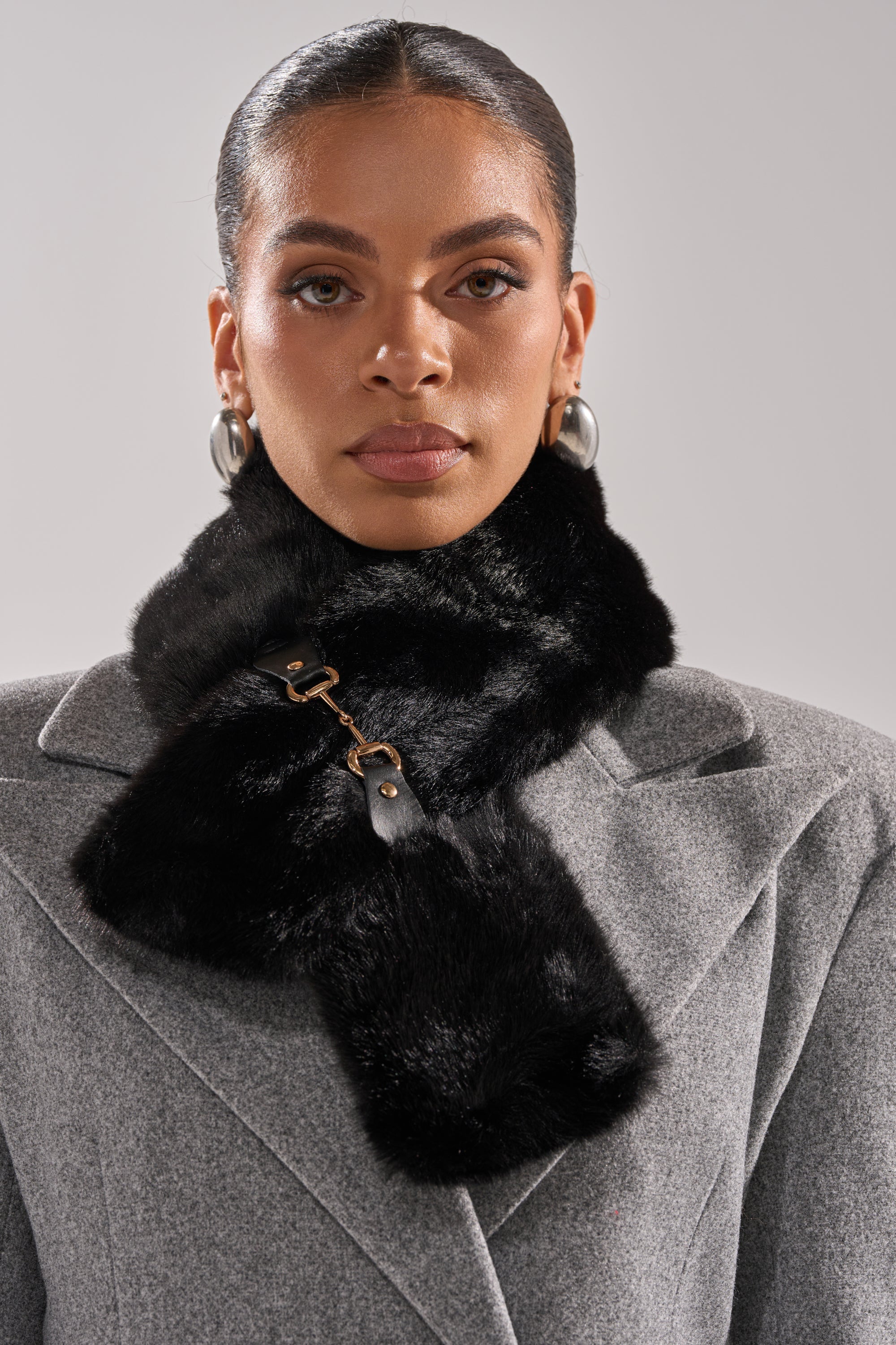 A woman in a grey wool coat wears the HORSEBIT FAUX FUR SCARF with a clasp, slicked-back hair, and large silver hoop earrings—an effortlessly chic look featuring a cozy accessory. 1