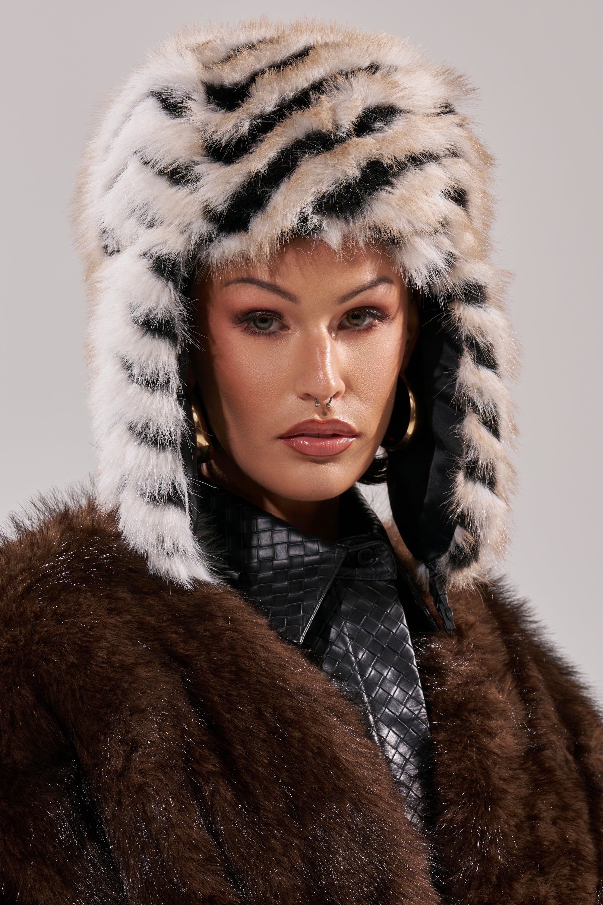 A person wearing the PORTIA AVIATOR HAT, a brown faux fur coat, black quilted shirt, and gold hoop earrings looks directly at the camera with a neutral expression. 9