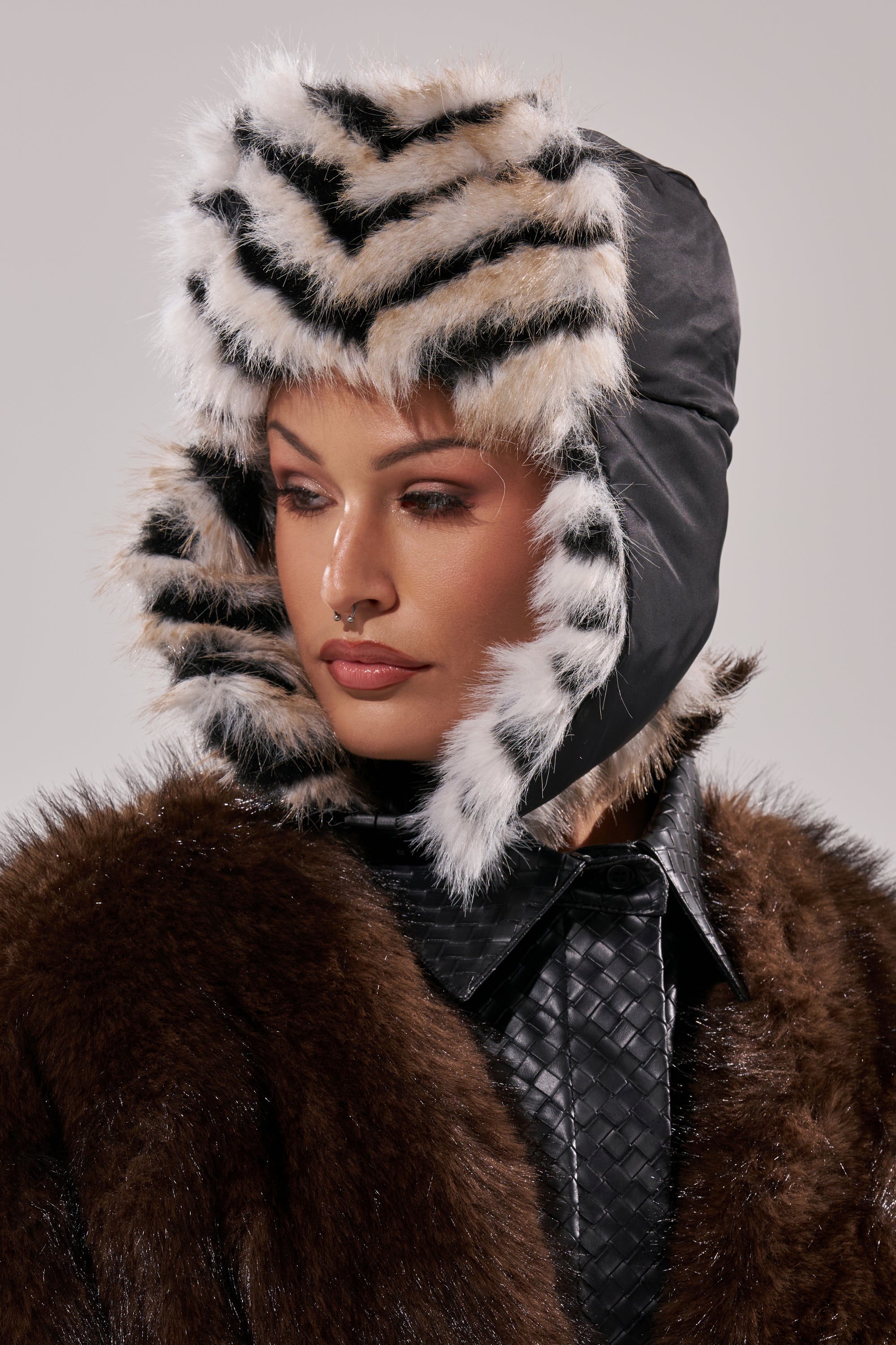 Wearing the PORTIA AVIATOR HAT in black and white faux fur with a chevron pattern, styled with a dark brown fur coat and black textured shirt, this individual glances to the side against a plain background. 15