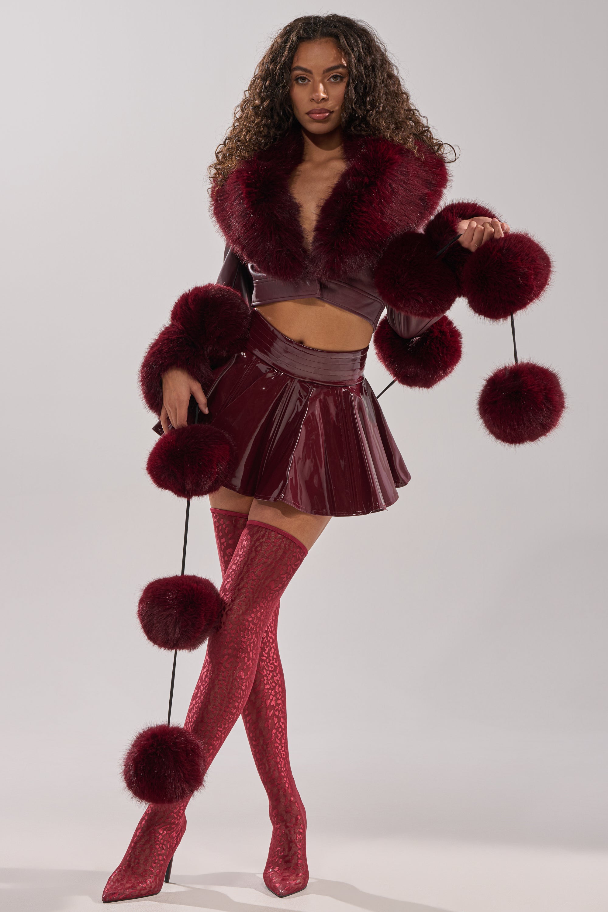 A woman stands against a plain background wearing a shiny burgundy crop top with fur trim, a matching mini skirt, patterned thigh-high boots, and holds the FEELIN BALLSY FAUX FUR SCARF adorned with oversized pom poms.