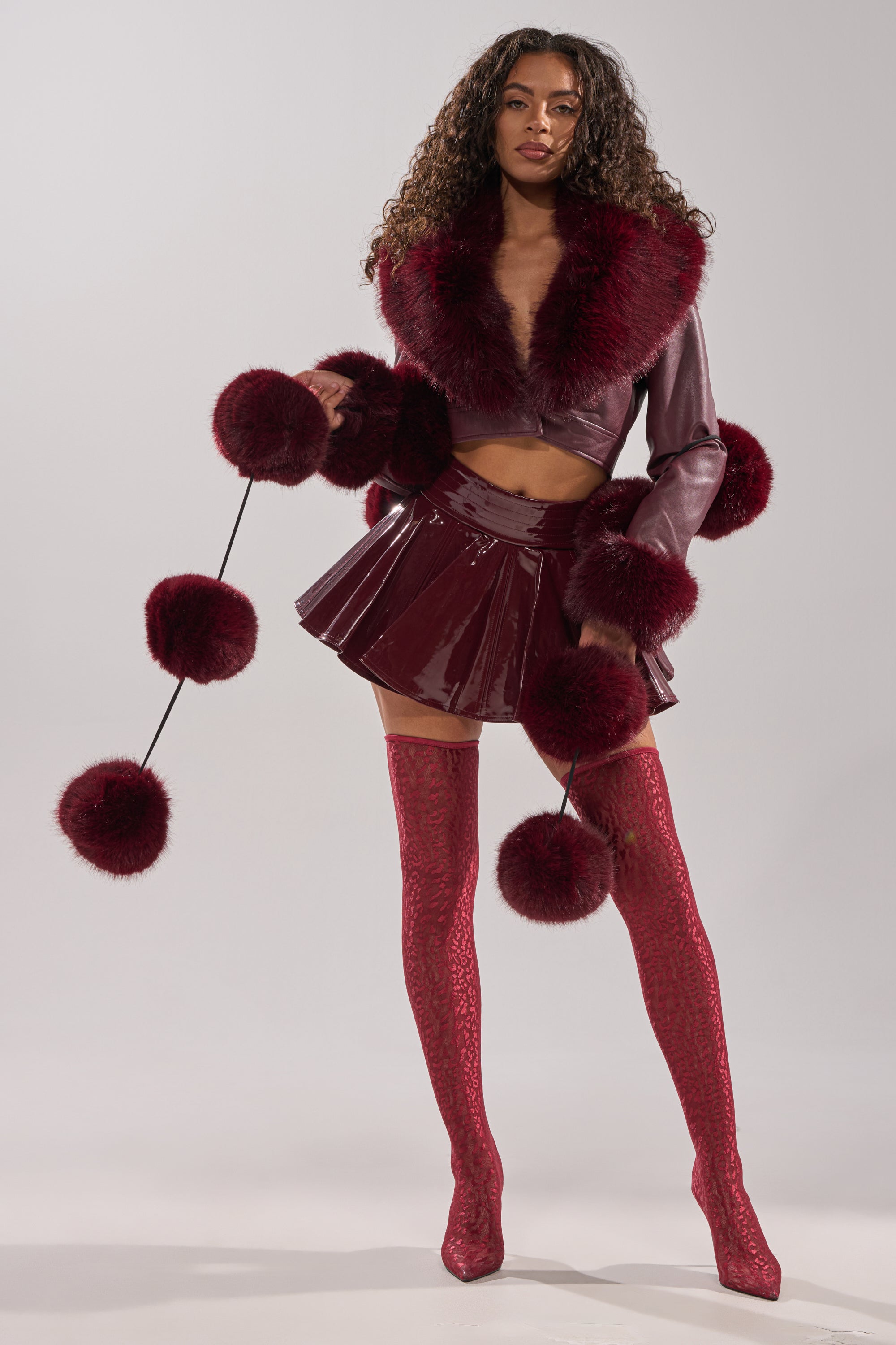 A woman poses confidently against a plain background, wearing a shiny burgundy mini skirt, matching crop jacket with large faux fur trim, red patterned thigh-high boots, and holds the FEELIN BALLSY FAUX FUR SCARF with oversized pom poms.