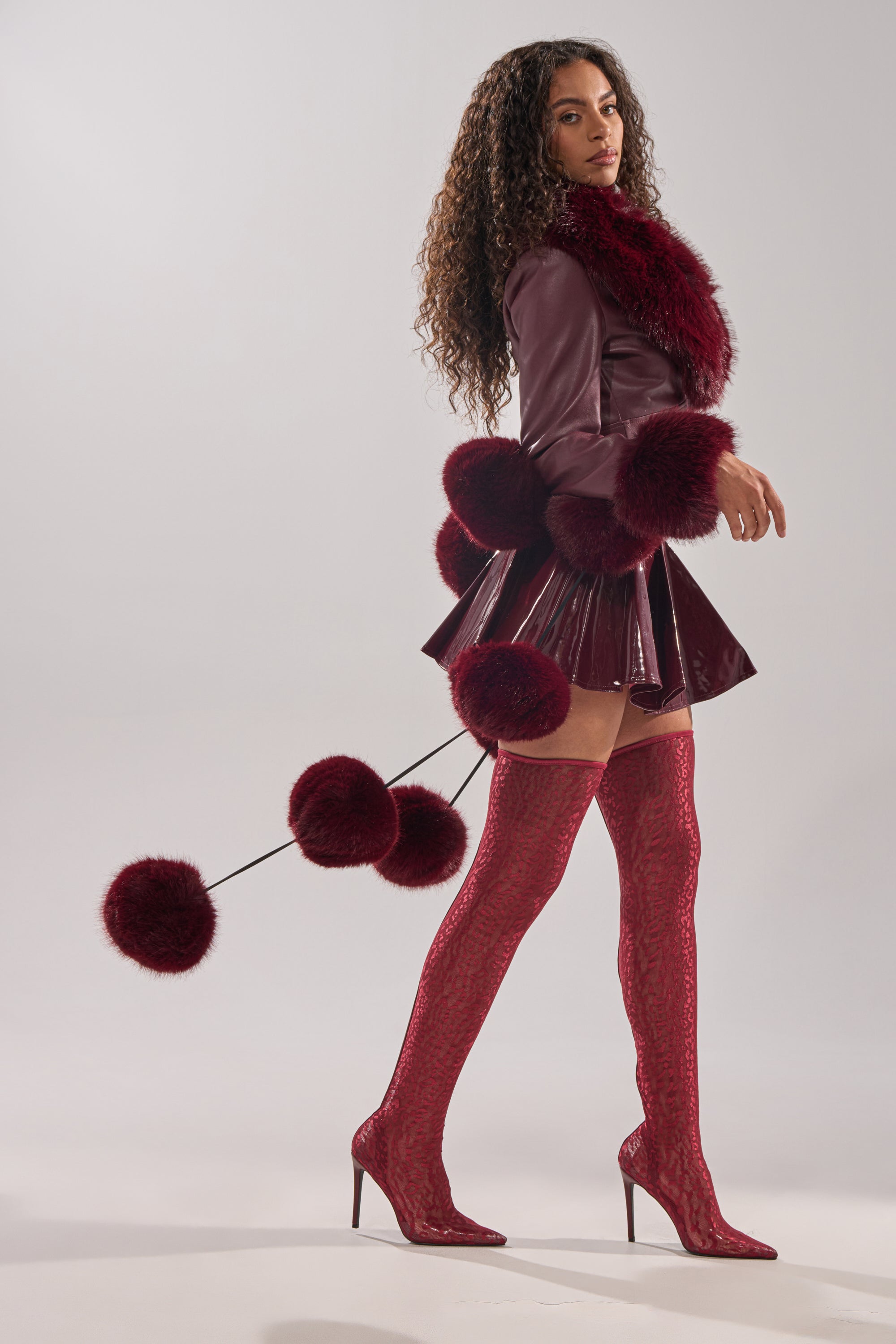 A woman with long curly hair models the FEELIN BALLSY FAUX FUR SCARF, styled with a maroon outfit featuring furry accents and oversized pom poms, paired with thigh-high patterned boots against a plain light background.