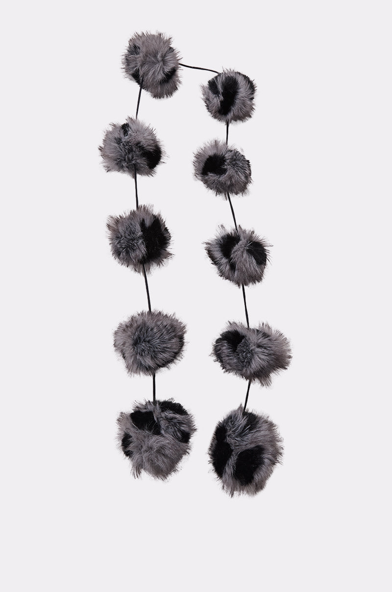 The BALLS TO THE WALL FAUX FUR SCARF features a black knit design with multiple oversized, fluffy black and gray faux fur pom-poms spaced along its length, displayed vertically against a plain light background.