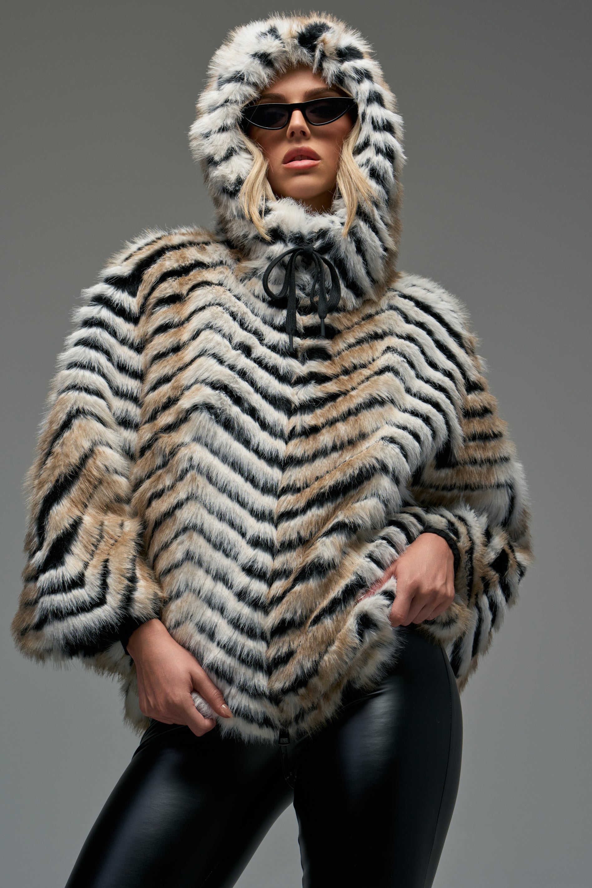 Draped in the CHEVRON DREAMING FAUX FUR STOLE, paired with black sunglasses and sleek black leather pants, a person strikes a confident pose against a plain gray background, perfectly capturing cozy-chic winter style.