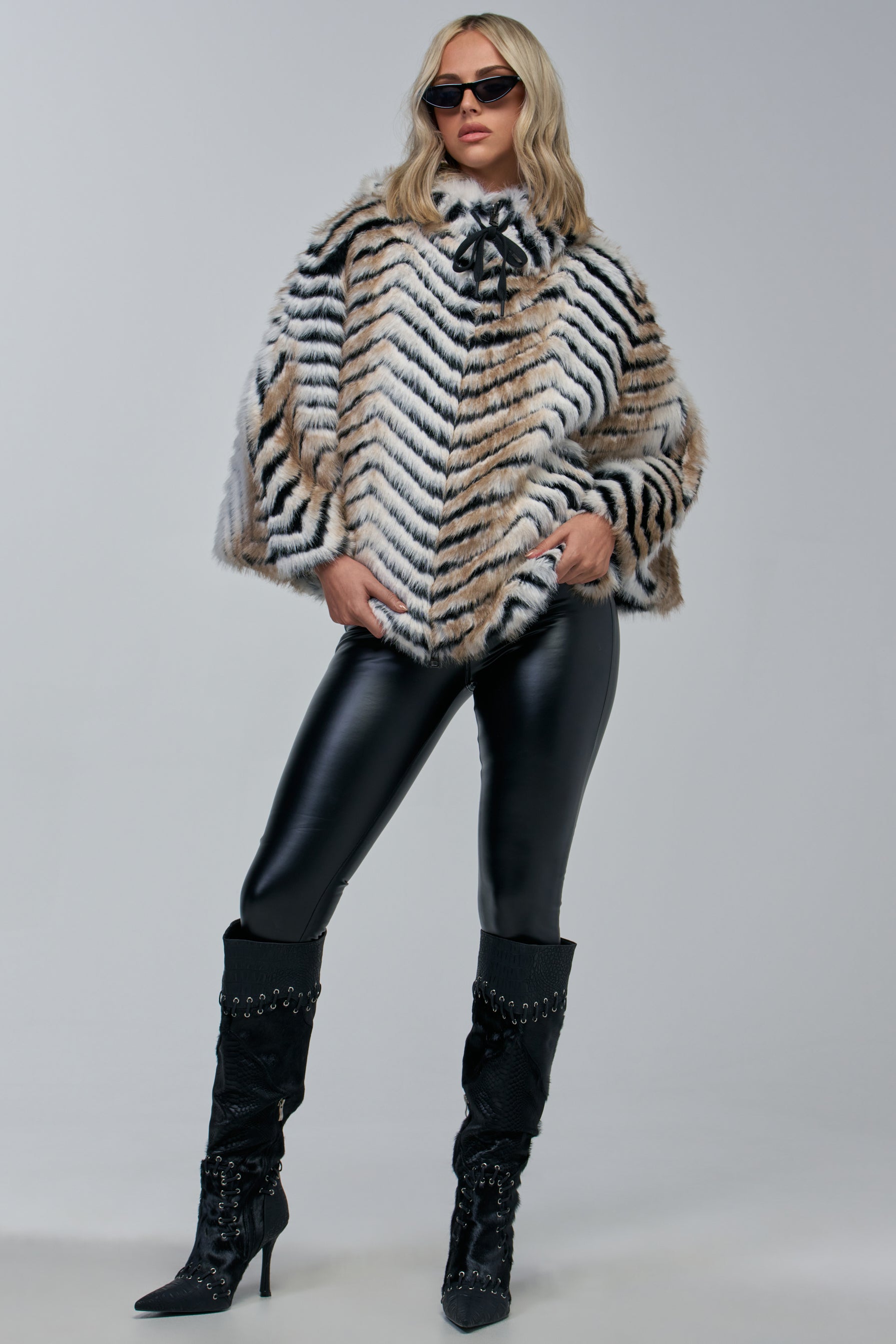 A woman stands confidently against a plain background, embodying effortless cozy-chic winter styling in the CHEVRON DREAMING FAUX FUR PONCHO, paired with sunglasses, black leather pants, and high-heeled black boots.