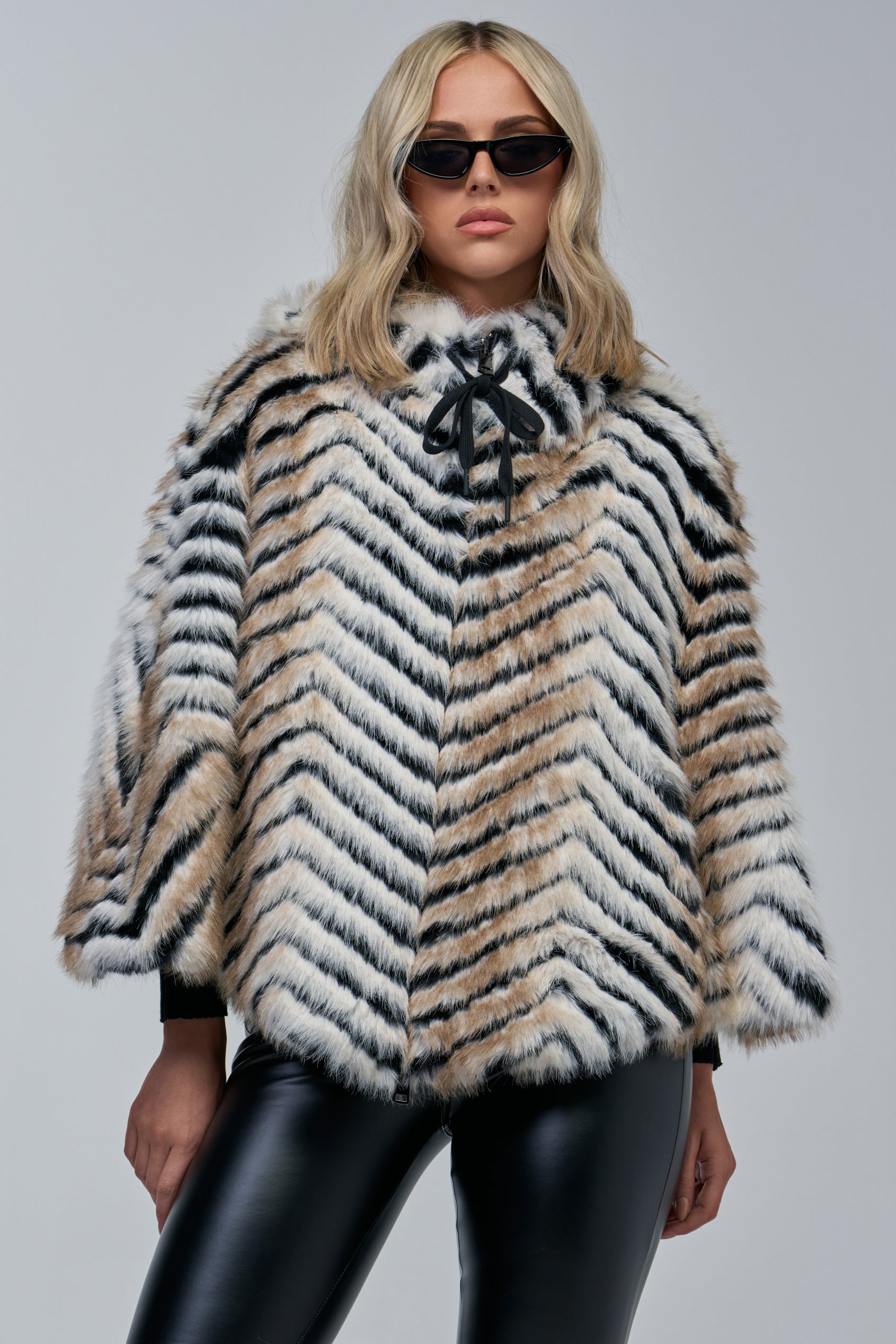 A person wearing black sunglasses, the CHEVRON DREAMING FAUX FUR PONCHO featuring cozy-chic black, white, and tan chevron stripes, and black leather pants stands against a plain gray background.