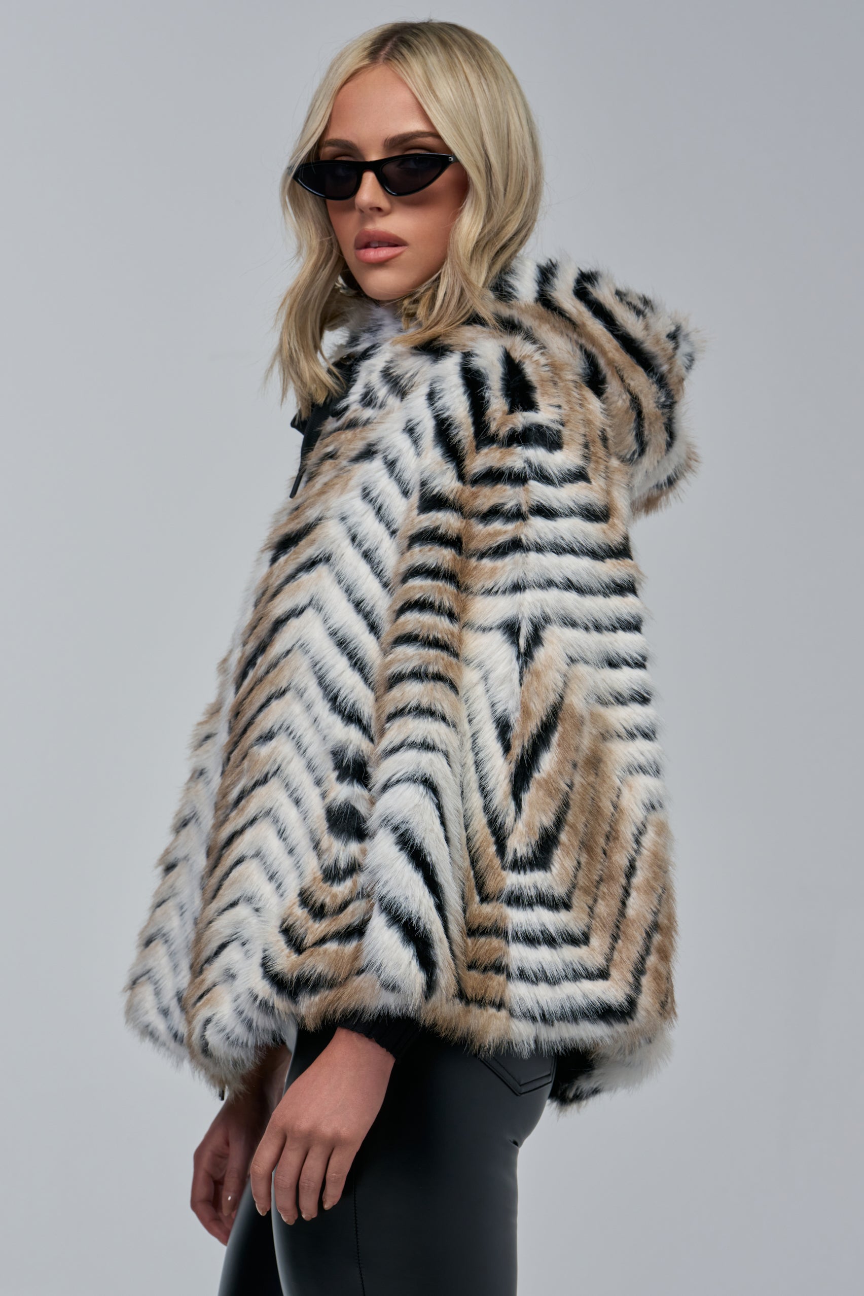 A woman wearing black sunglasses and the CHEVRON DREAMING FAUX FUR PONCHO with a bold tiger-stripe chevron pattern stands against a gray background, looking over her shoulder—a perfect example of cozy-chic winter styling.