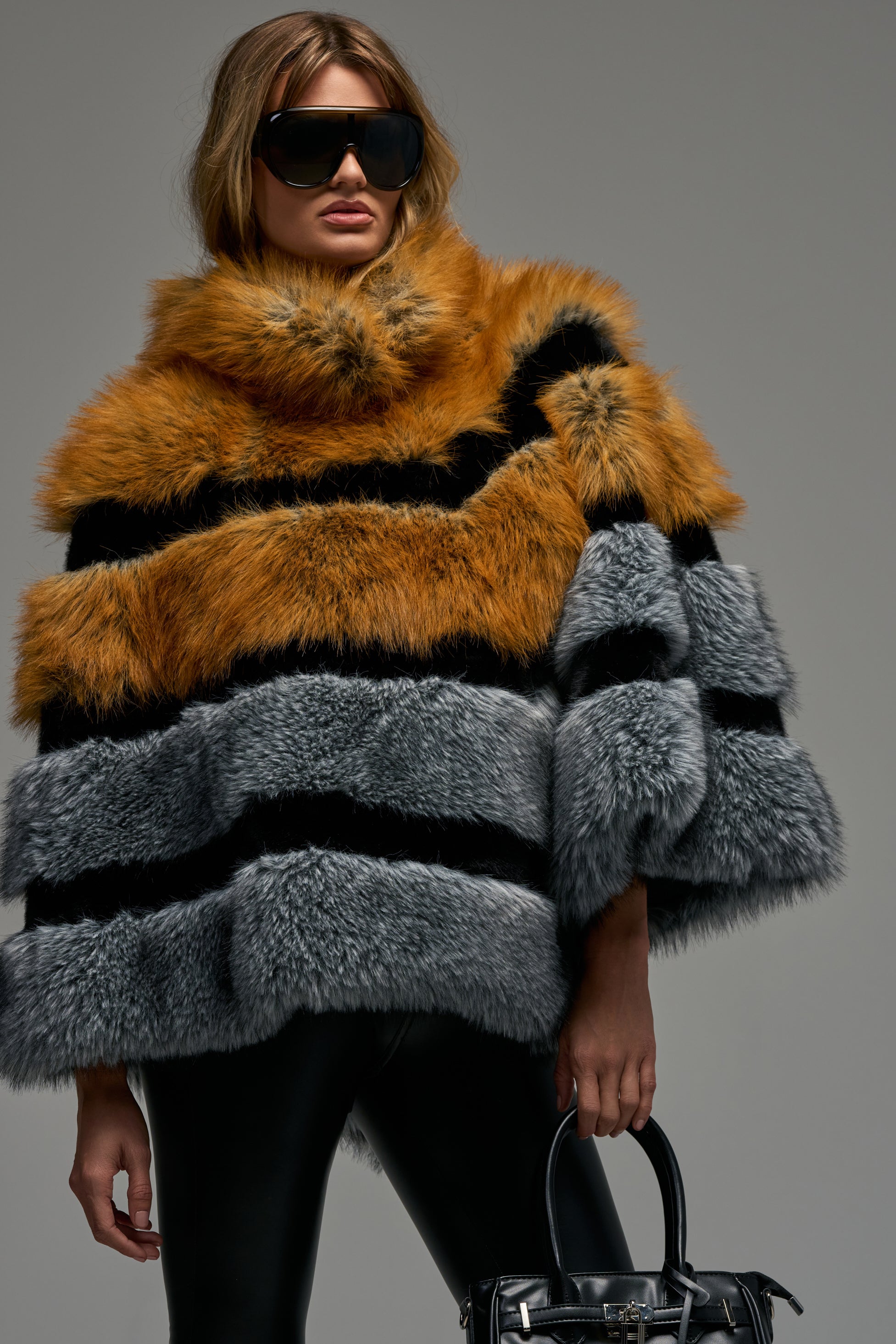 A person wearing oversized sunglasses and the VINTAGE VIBES FAUX FUR STOLE—a luxe cold weather accessory with orange, black, and gray stripes—holds a black handbag while standing against a plain gray background.