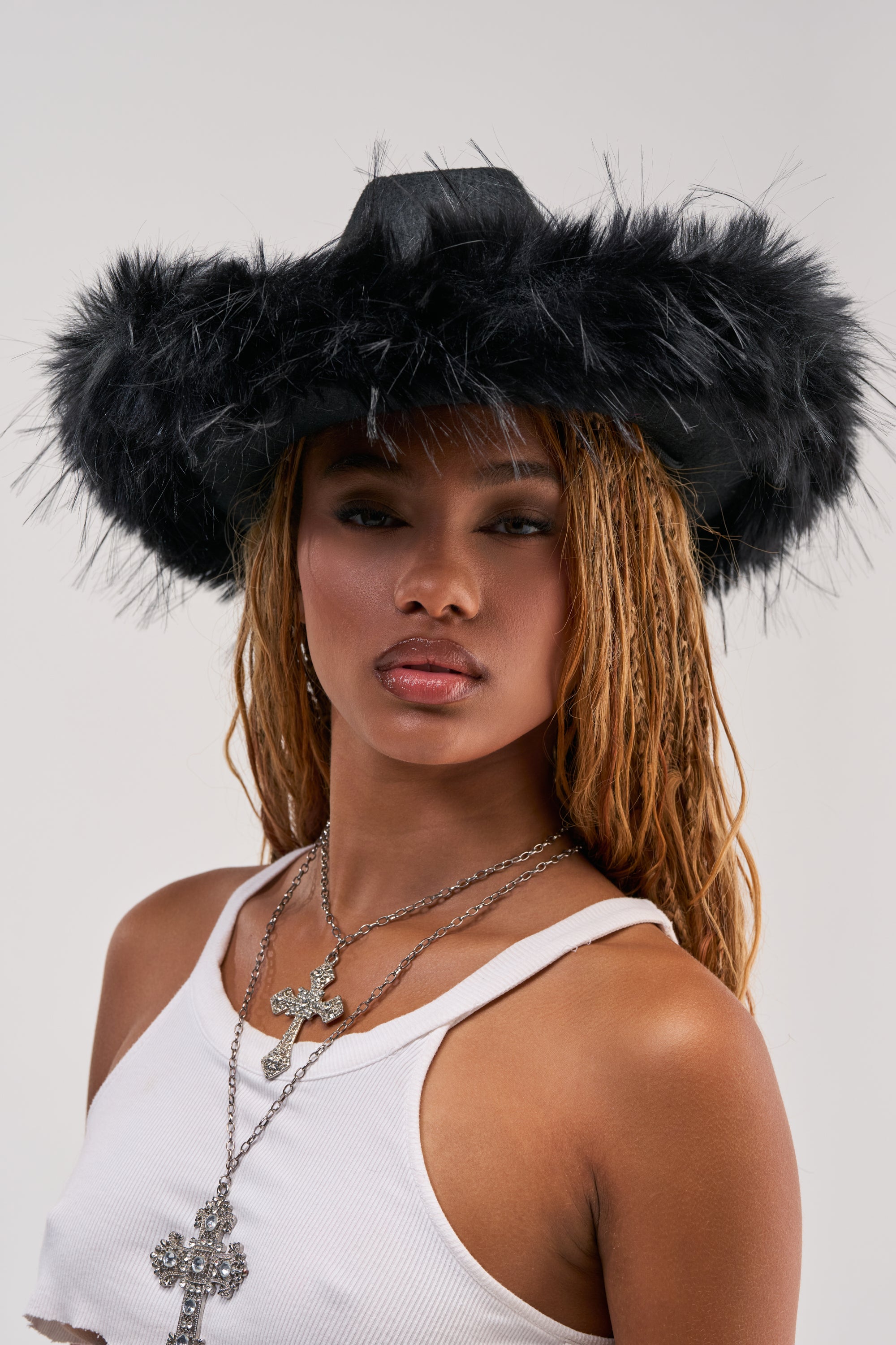 A woman poses against a plain light background, showcasing the SADDLE UP FUR TRIM HAT—a bold black cowboy hat with plush fur trim. Styled with a white tank top and layered silver cross necklaces, her long hair and neutral expression complete this modern western look, perfect for festival season.