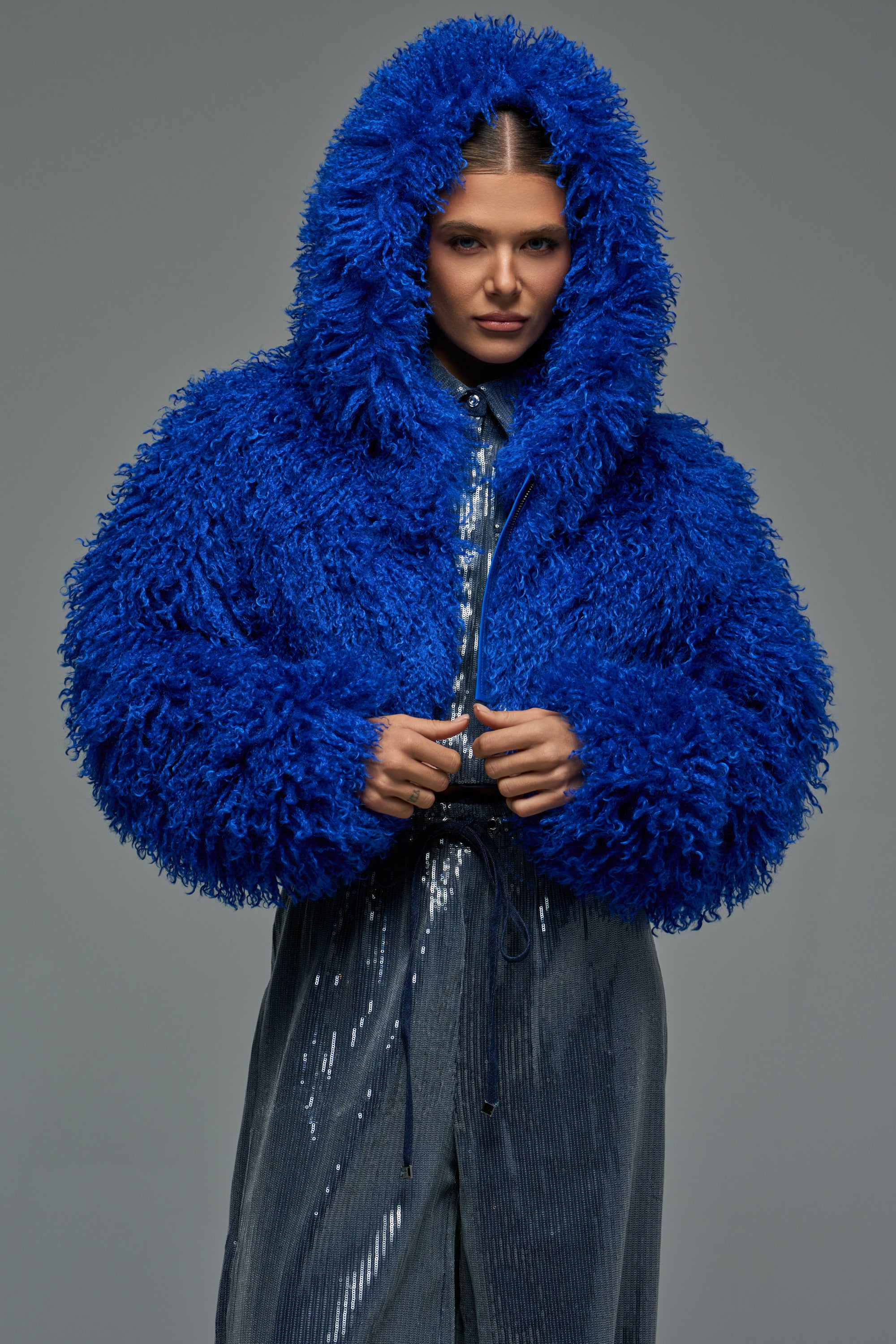 A person wearing an ELECTRIC LOVE FAUX FUR BOMBER, a cropped, electric blue faux fur jacket with a large hood.