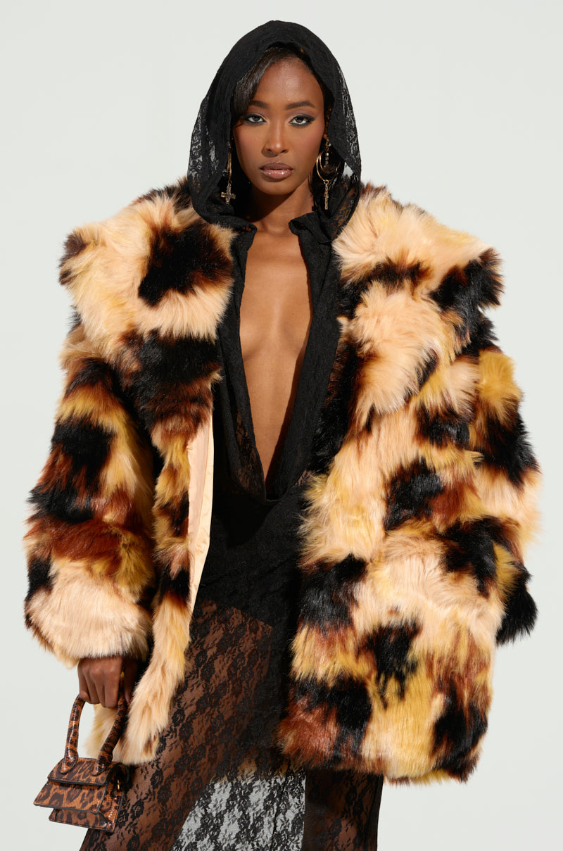Draped in the ARI OVERSIZED FUR COAT, a woman exudes confidence as the coat's vibrant hues beautifully contrast with her sleek black hooded ensemble underneath. She accessorizes with a small purse and eye-catching statement earrings, all set against a simple backdrop.