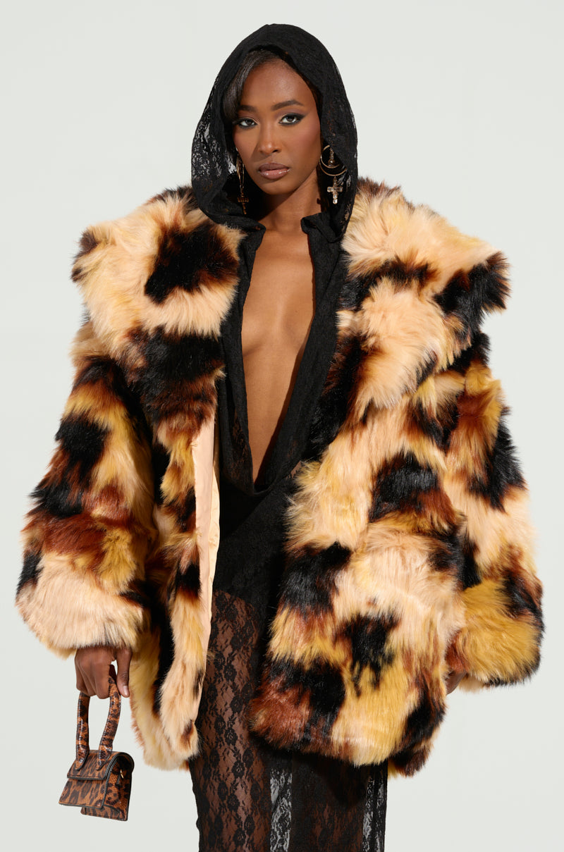 A person wearing the ARI OVERSIZED FUR COAT with a black hooded garment underneath is holding a small leopard-print handbag. The plain, light-colored background emphasizes their bold fashion choice.