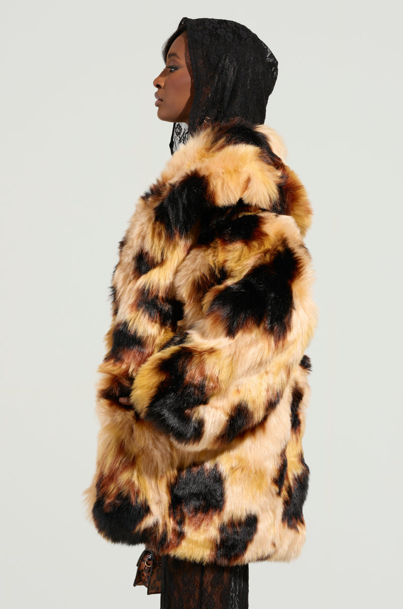 A person stands in profile wearing the ARI OVERSIZED FUR COAT, which features a luxurious blend of brown, black, and beige faux fur. The coat includes a satin-lined interior for added elegance. They also wear a black lace head covering against the plain light gray background.