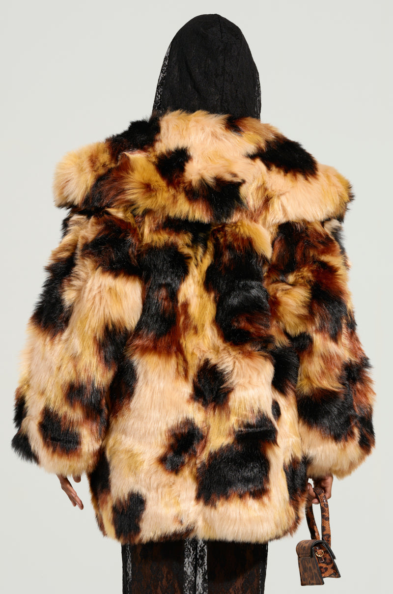A person wearing the ARI OVERSIZED FUR COAT, featuring luxurious black, brown, and beige patches, stands with their back to the camera. The satin-lined interior is visible as they hold a small brown handbag and wear a black hood beneath the coat's large collar.