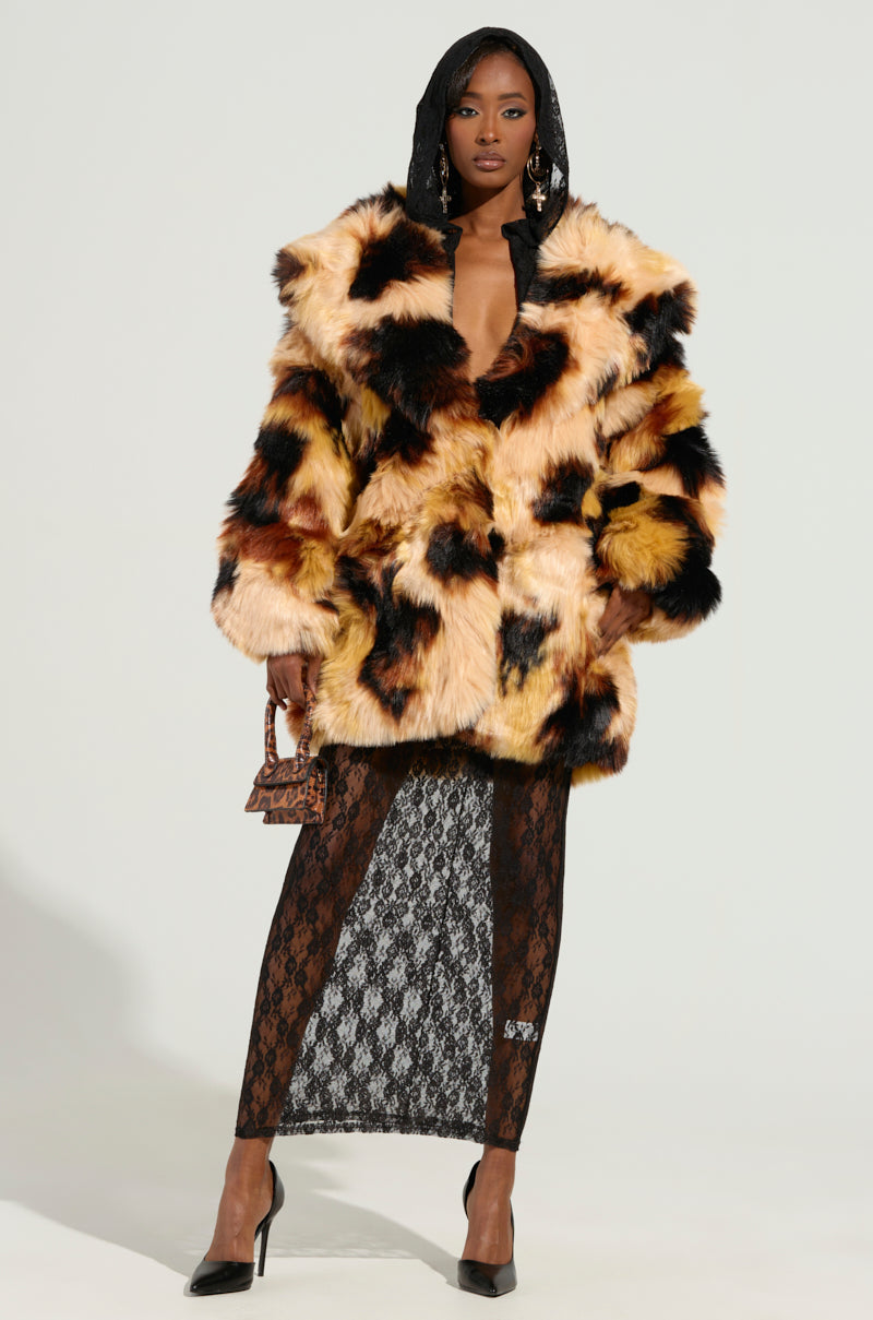 Dressed confidently, a person showcases the ARI OVERSIZED FUR COAT, featuring an oversized design with a satin-lined interior in shades of black, brown, and tan. They wear it over a lace dress while holding a small handbag and wearing black high heels, posing elegantly against a plain background.