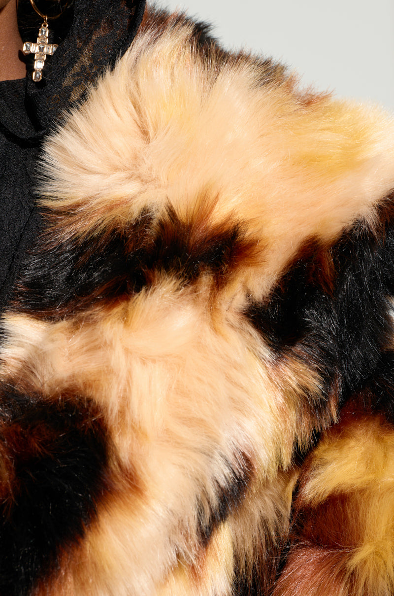 Close-up of the vibrant ARI OVERSIZED FUR COAT featuring an array of brown, black, beige, and cream tones. A glimpse of a black garment can be seen beneath it, accompanied by an earring embellished with small pearls and a cross charm.
