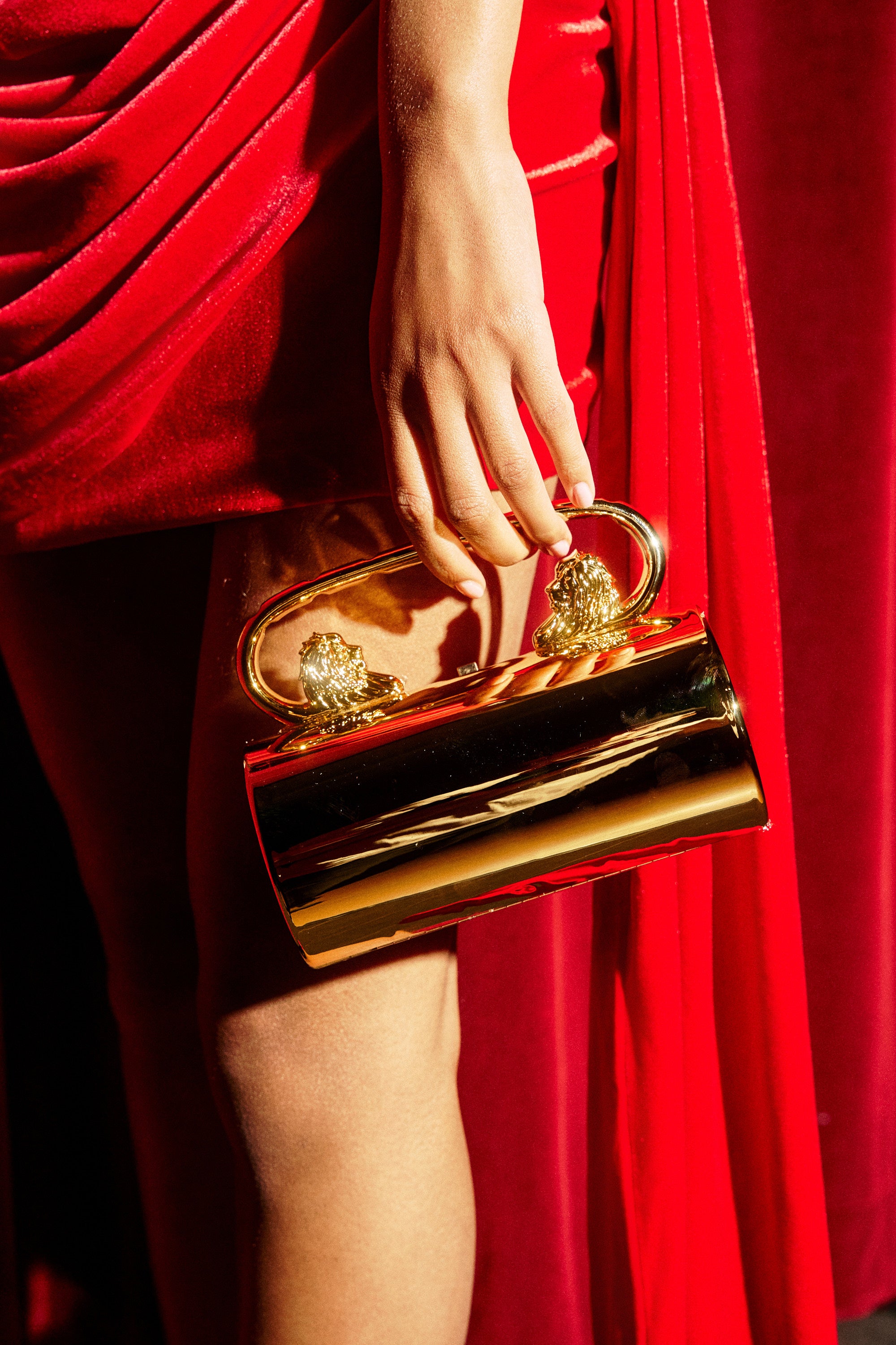 Holding the LETS GO OUT CLUTCH BAG with two metallic handles, a person in a red dress poses against a red velvet background.