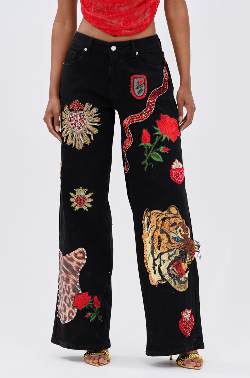 A person wears FIERCE DENIM PANT, a black high-rise fit with bold embroidered patches—tiger, snake, leopard print, roses, and hearts—paired with a red crop top and open-toe heels.