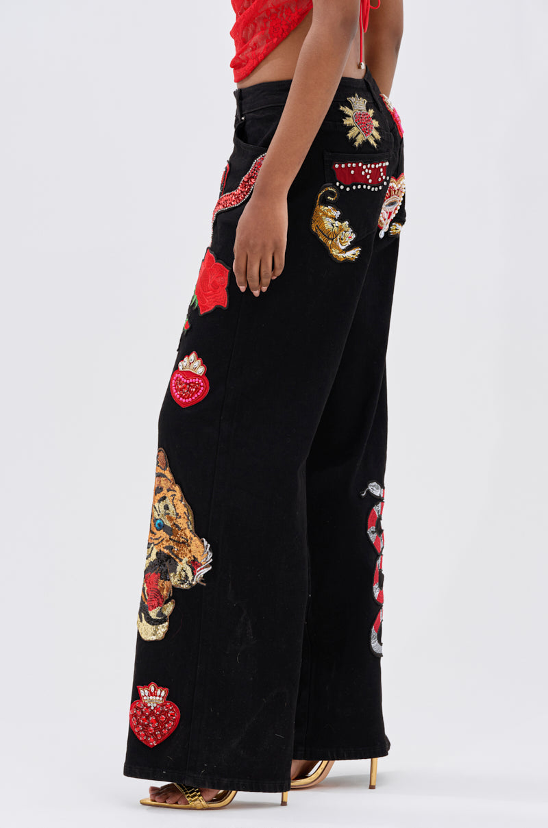A person wears the FIERCE DENIM PANT—black wide-leg jeans with bold embroidered patches—styled with a red lace crop top and gold heels for a rebellious glam look.