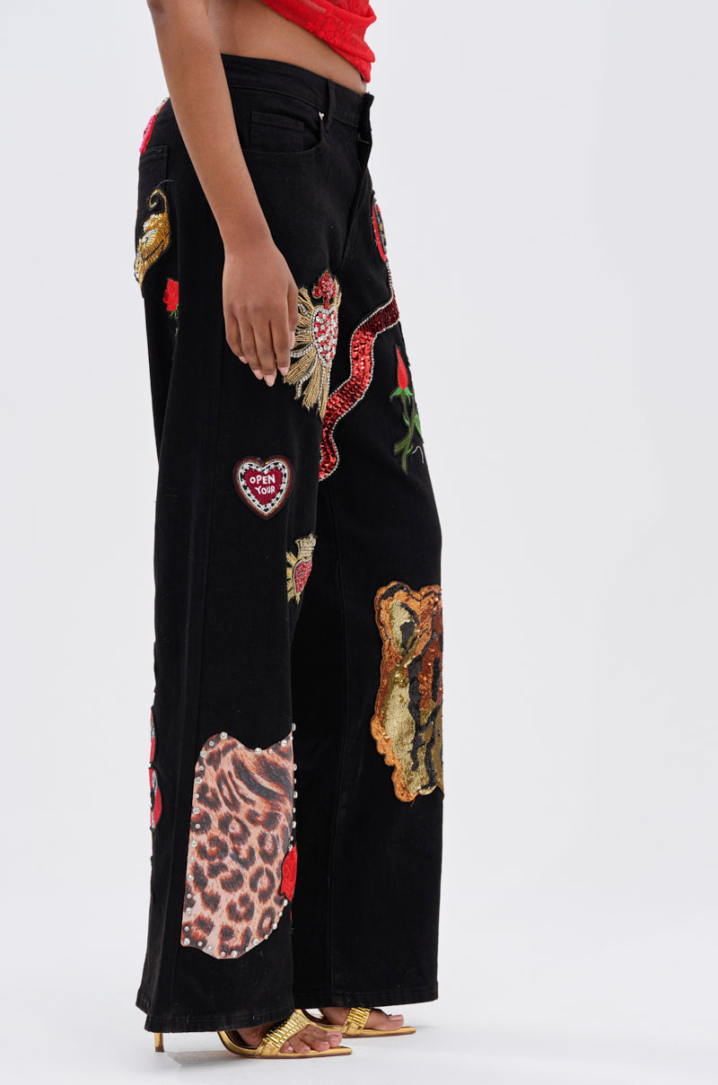 Someone rocks the FIERCE DENIM PANT—black wide-leg jeans adorned with colorful embroidered patches, including a red snake, leopard print, and a heart—styled with a red top and gold heeled sandals.