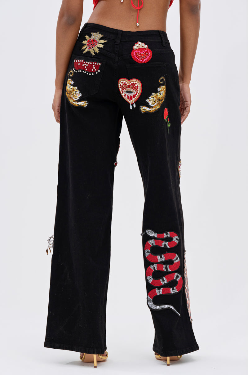 Wearing the FIERCE DENIM PANT—black wide-leg jeans with embroidered and beaded details like a snake, hearts, wings, and a rose—a person stands in heels facing away, radiating rebellious glam.