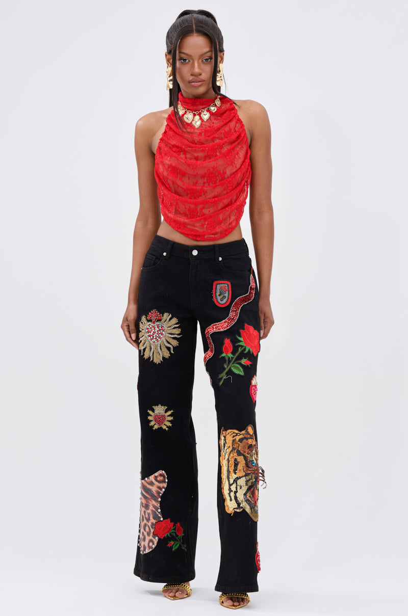 A woman stands facing forward in a sleeveless red draped top and the FIERCE DENIM PANT, featuring embroidered flower, snake, leopard, and tiger patches. With long hair and gold accessories, she exudes rebellious glam.