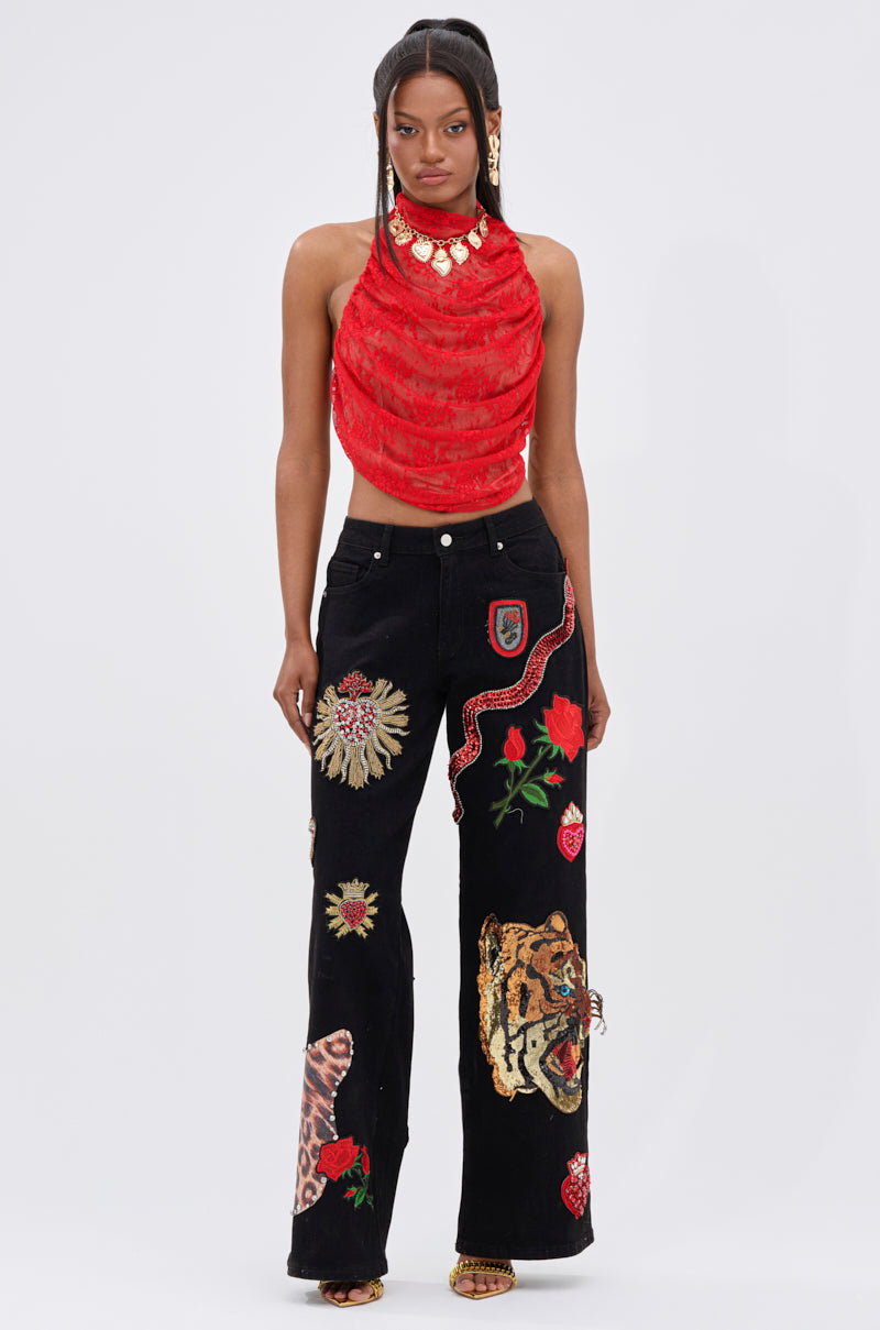 A woman faces forward in a sleeveless red draped top and FIERCE DENIM PANT adorned with colorful embroidered patches—tiger, snake, flowers, and leopard. Gold jewelry and sleek straight hair complete her rebellious glam look.