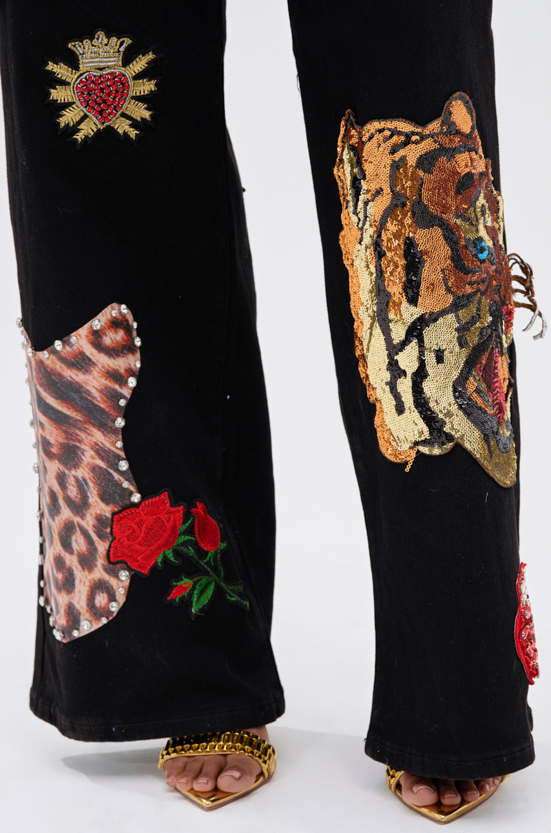 The FIERCE DENIM PANT stands out with embroidered patches—crowned heart, tiger face, roses, animal print, and metallic studs. Styled with open-toe gold heels, this rebellious glam look pops against a plain white background.
