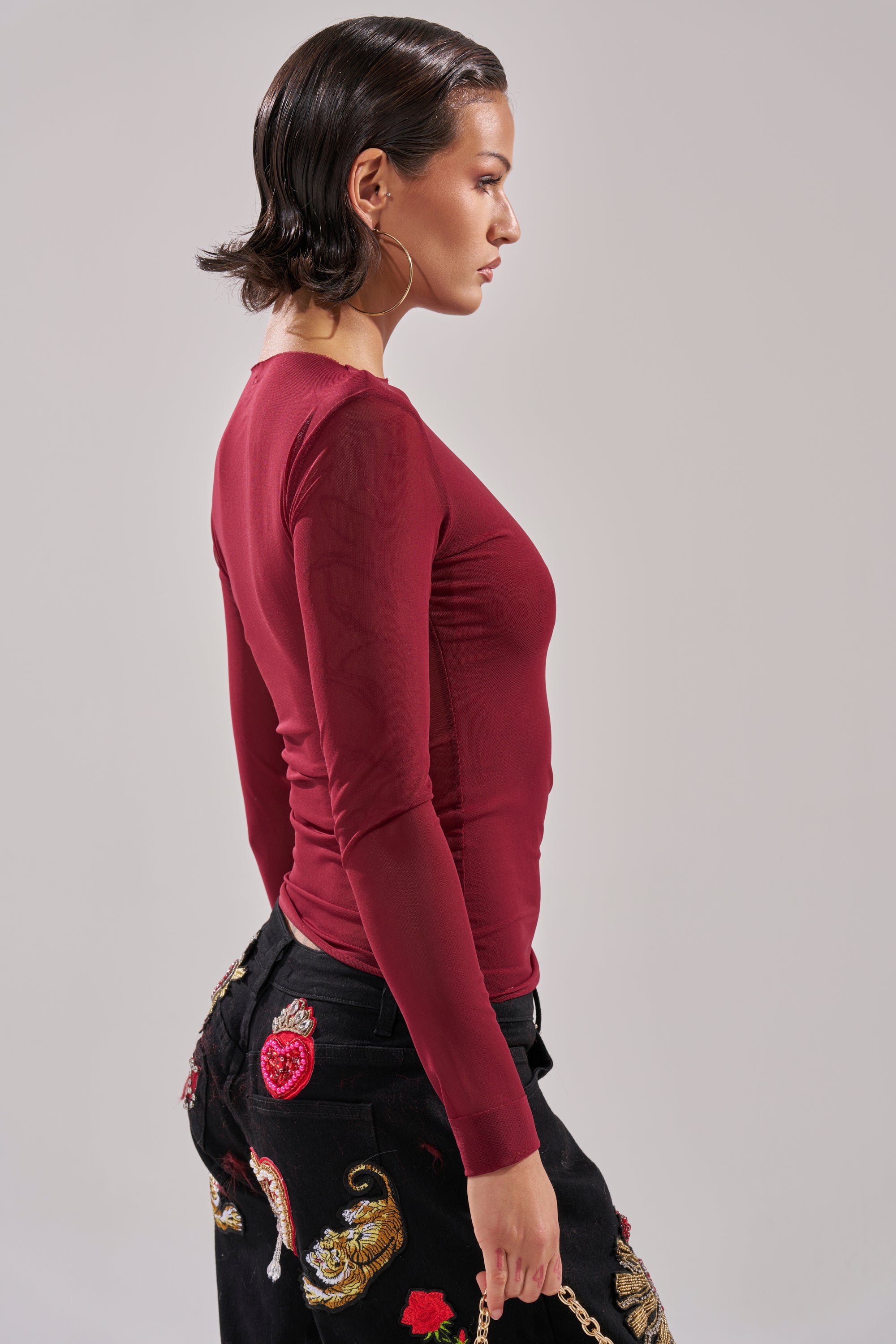 A person with short dark hair wears the PAXTON Double Lined Mesh Long Sleeve Top in plum and black pants with colorful embroidery, shown in profile against a plain light background.
