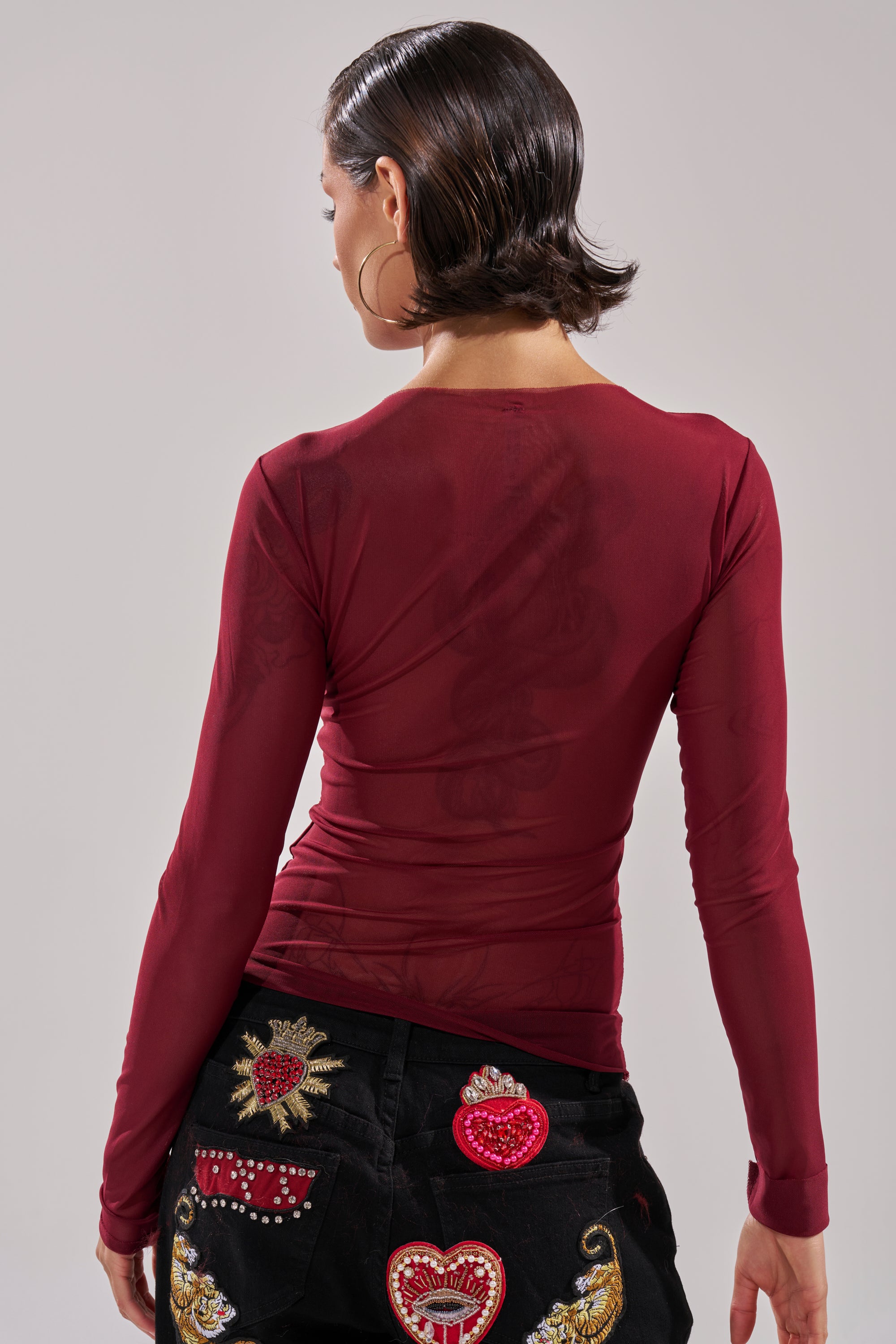 A person with short, slicked-back dark hair wears the PAXTON DOUBLE LINED MESH LONG SLEEVE TOP IN PLUM and black jeans with colorful embroidered heart patches, facing away from the camera against a neutral background.