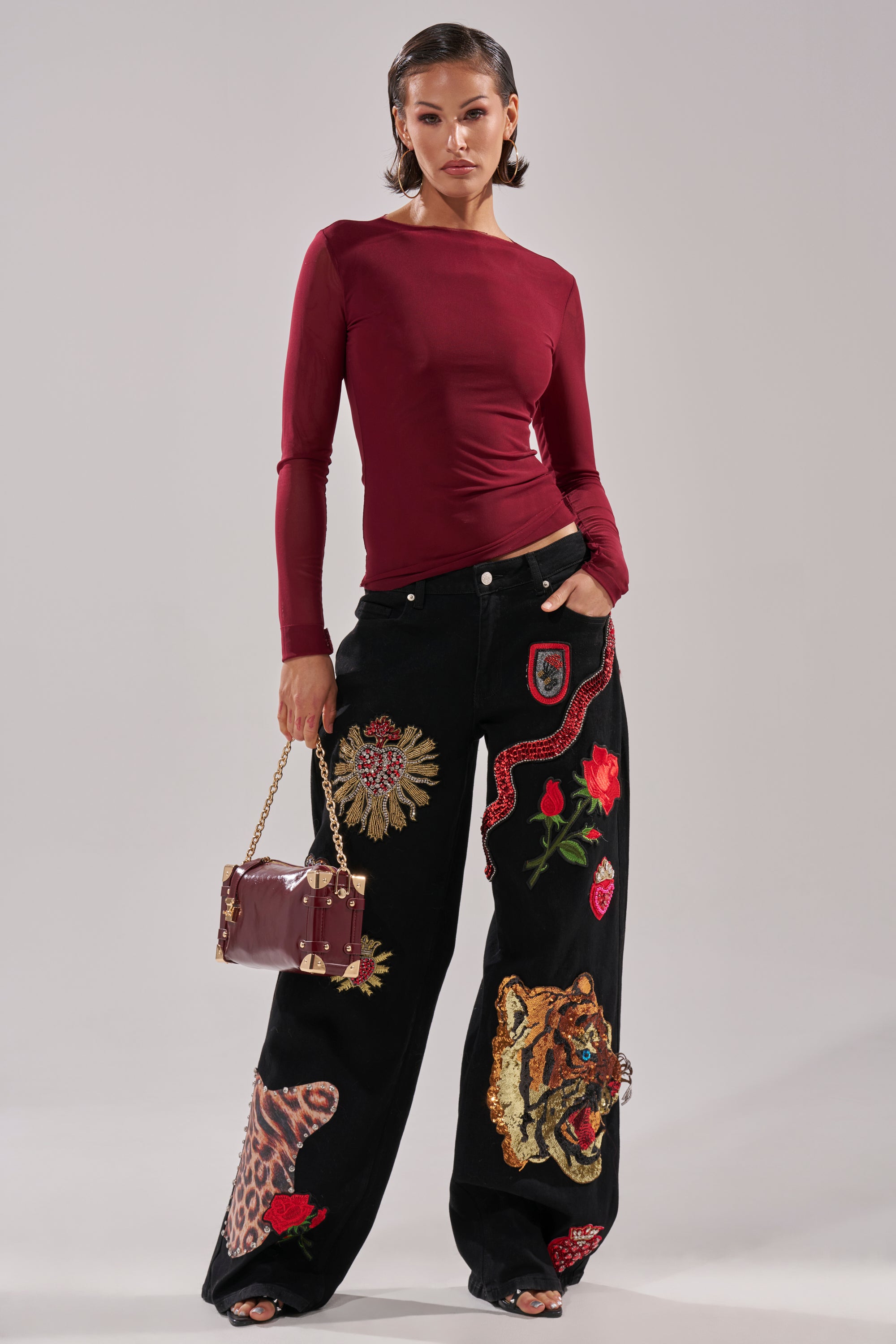 A woman stands confidently in the PAXTON Double Lined Mesh Long Sleeve Top in Plum, paired with high-waisted black jeans with bold animal and floral embroidery. She carries a burgundy handbag and styles her dark hair slicked back.