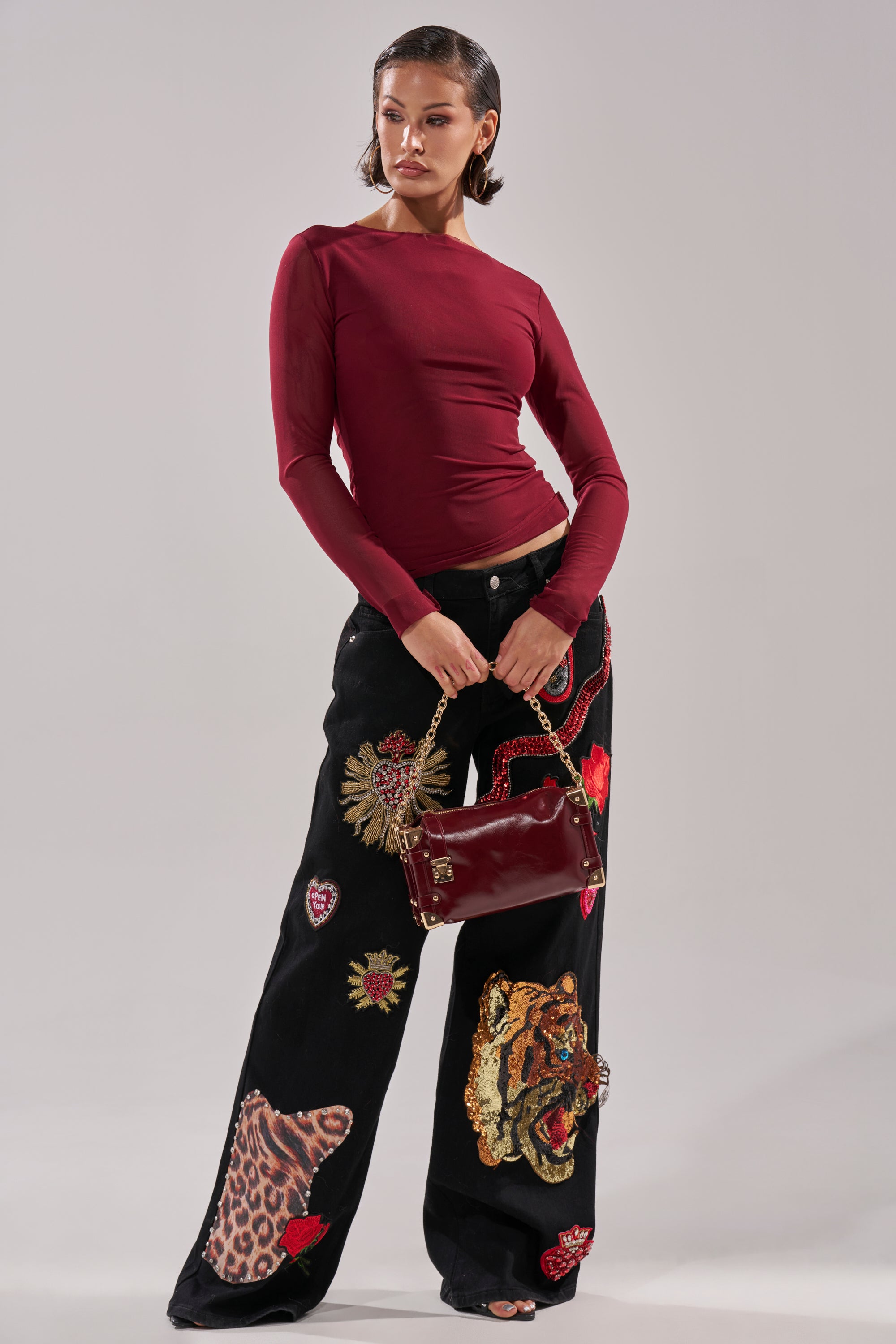 A woman poses in the PAXTON Double Lined Mesh Long Sleeve Top in Plum with black wide-leg pants featuring colorful embroidered animals, holding a shiny burgundy handbag against a plain light background.